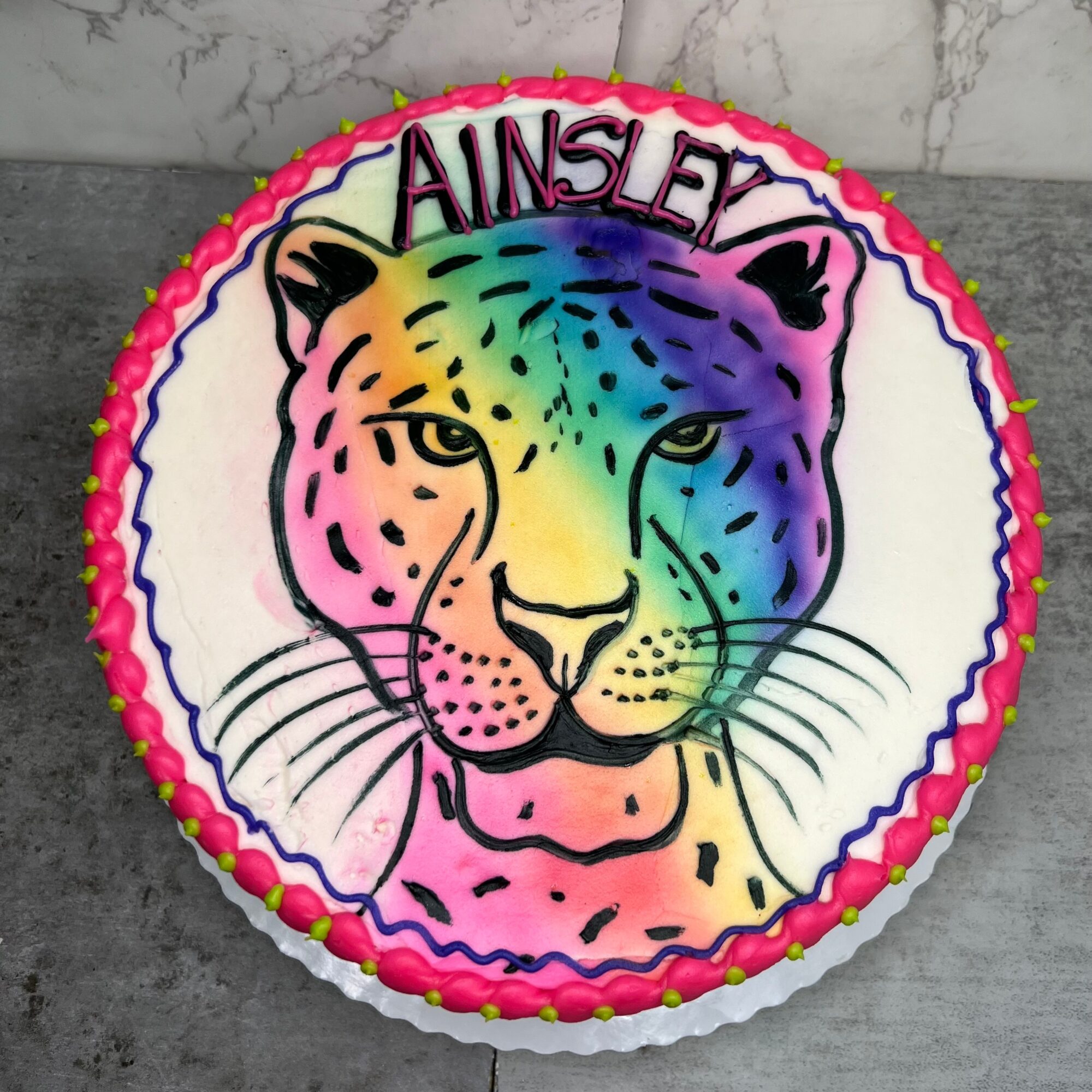 Colorful leopard face with the name 'Ainsley' above, decorated with pink, yellow, green, blue, and purple colors, on a birthday cake.