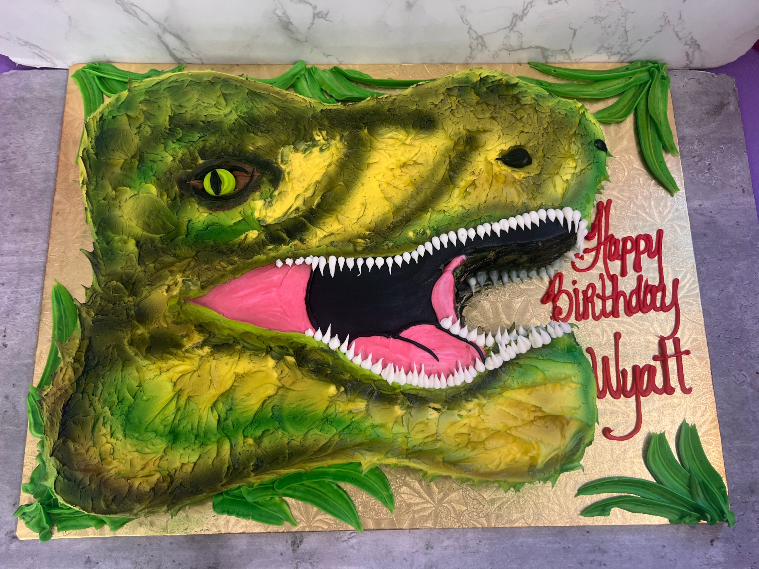 Birthday cake decorated as a crocodile head with green icing, yellow eyes, sharp teeth, and pink tongue, with green leaves around edges.