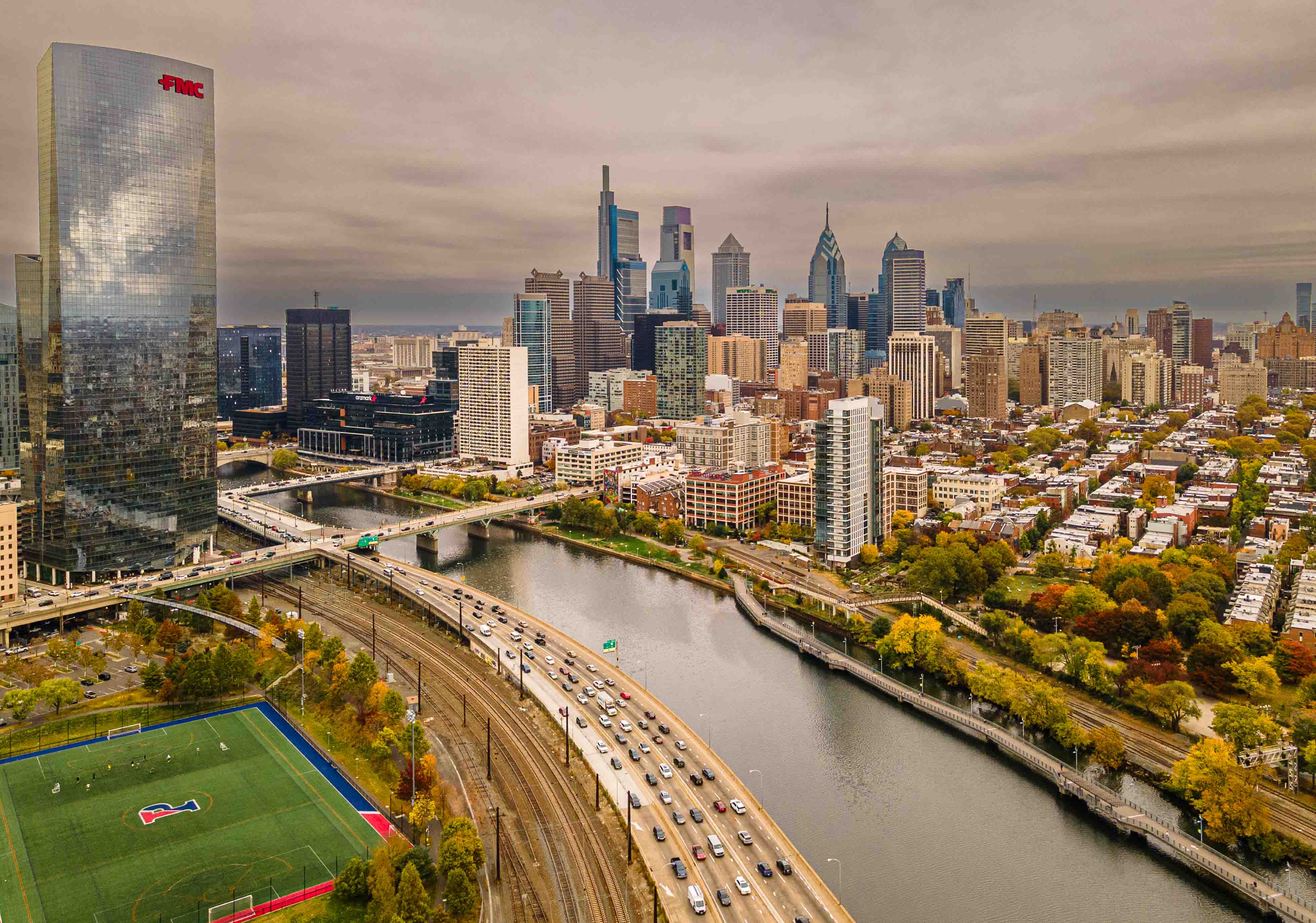 City skyline with tall buildings, a river, a highway, and a sports field, under cloudy sky.
