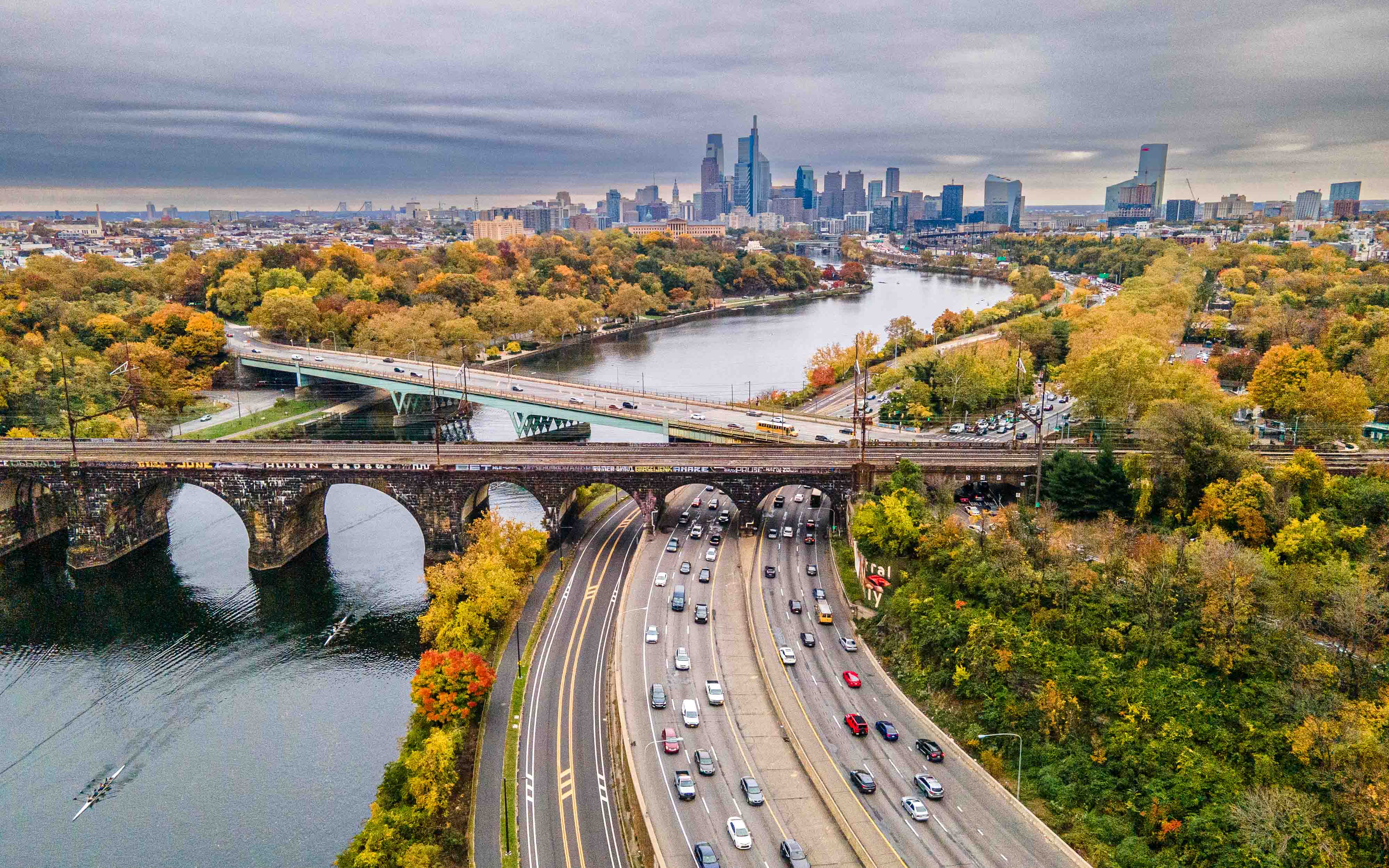City skyline with river, bridges, and highway surrounded by trees with autumn foliage, under cloudy sky.