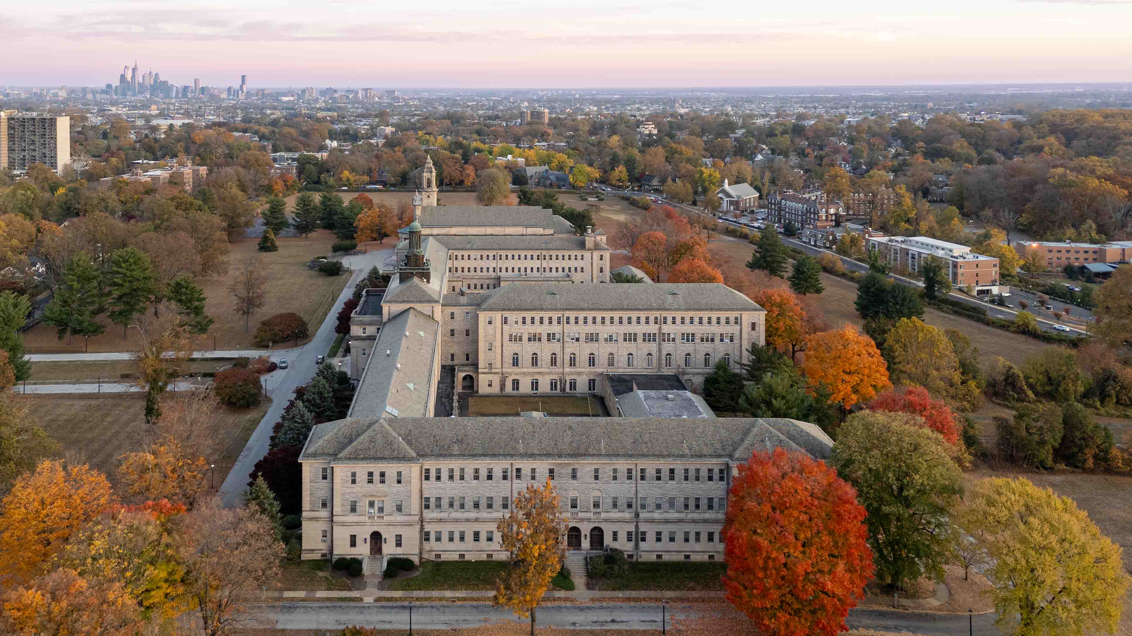 Aerial view of a large building complex surrounded by trees with autumn foliage, city skyline in the distance.