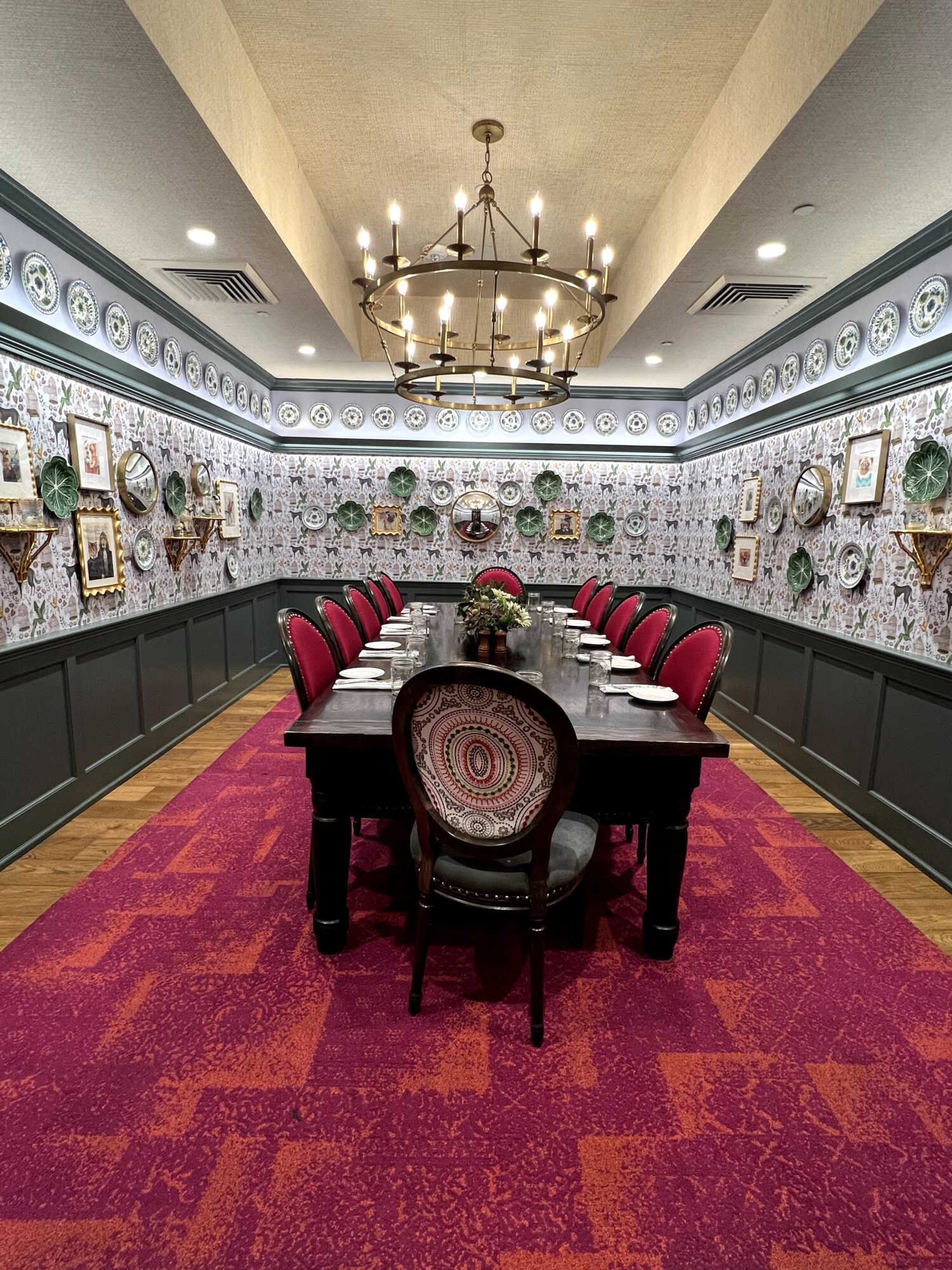 Long dining table with red chairs in a decorated room with framed pictures and a chandelier, carpeted floor, and ornate wall trim.