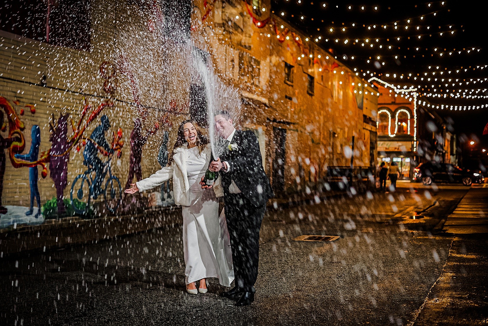 Two people in formal attire celebrating with a water spray on a decorated street at night.