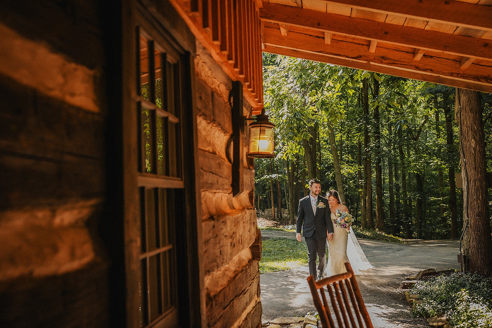 A couple walking outdoors near a wooden building with trees in the background, sunlight filtering through the leaves.