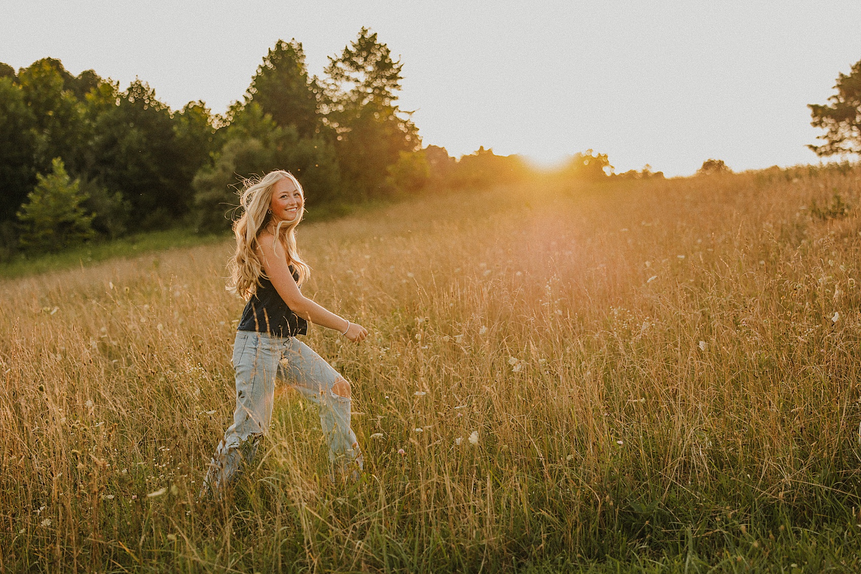 A girl walking through a grassy field at sunset, with trees in the background and sunlight shining behind her.