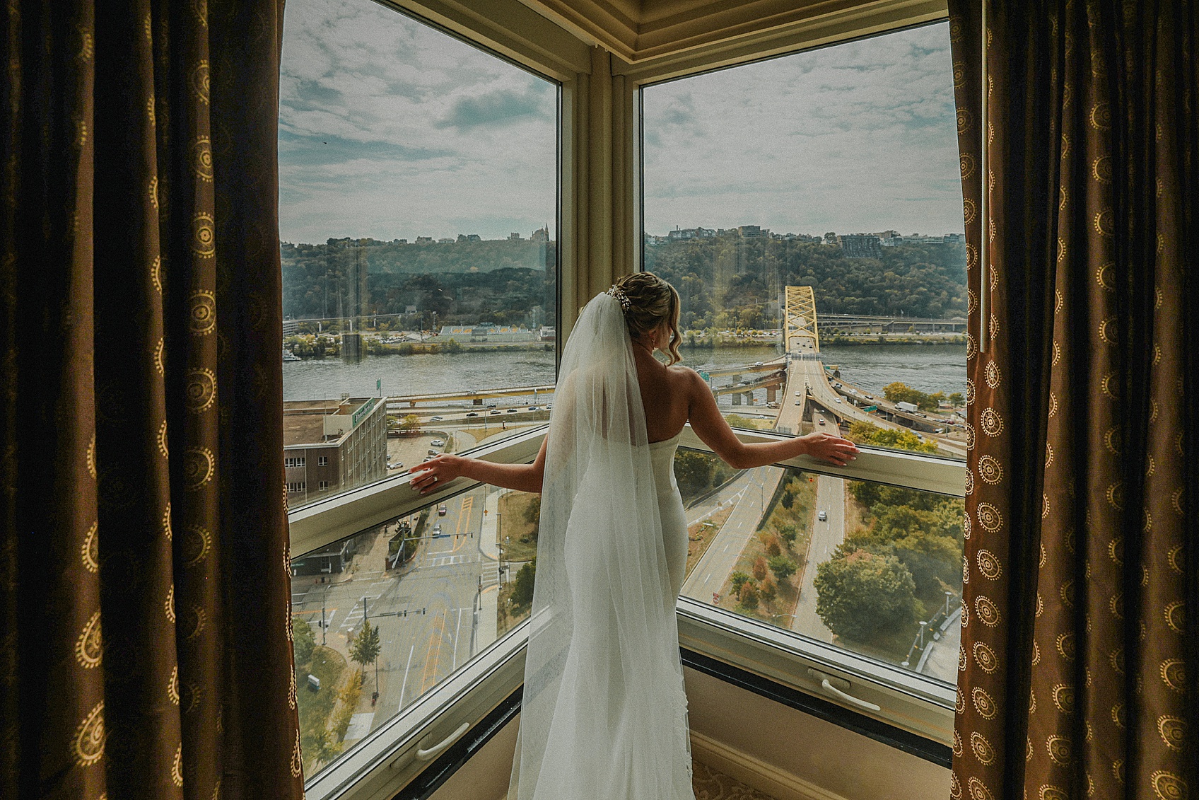 Woman in wedding dress and veil looking out of large corner window with city view.