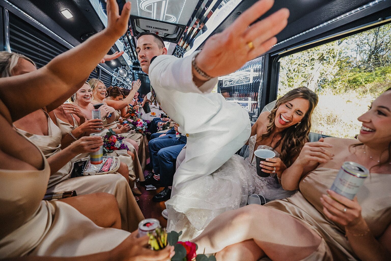 People celebrating on a bus, smiling, raising hands, and holding drinks, with sunlight outside the window.