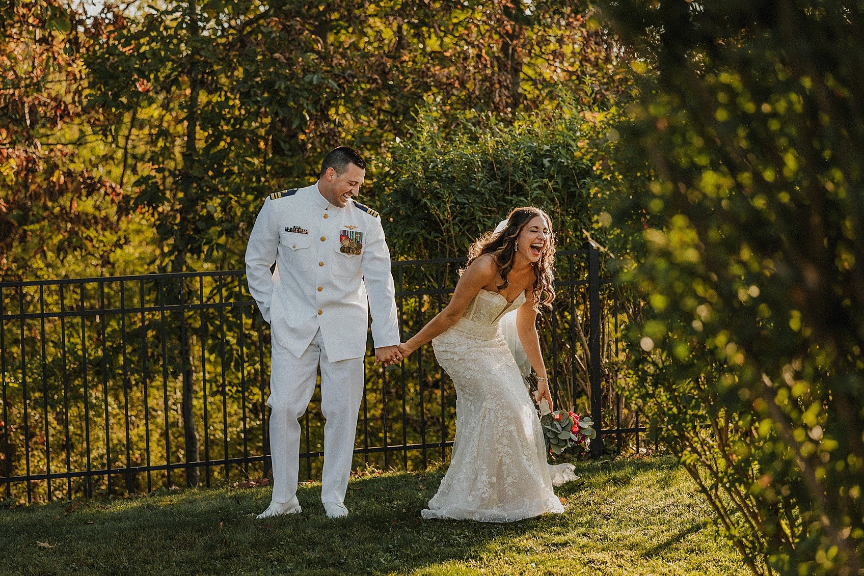 A bride and groom holding hands outdoors, with trees and a fence in the background, during daytime. The bride is smiling.