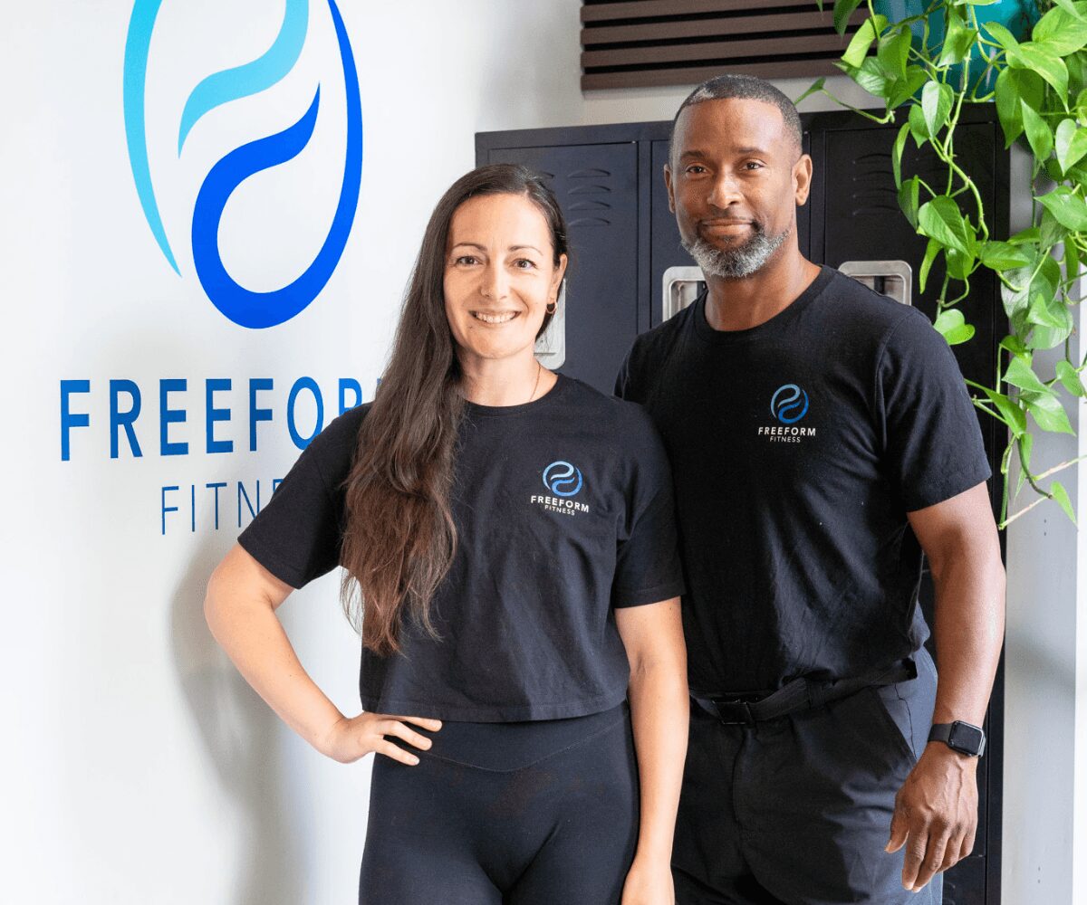 Two people standing indoors, smiling, wearing black t-shirts with a logo, in front of a wall with a logo and a plant.