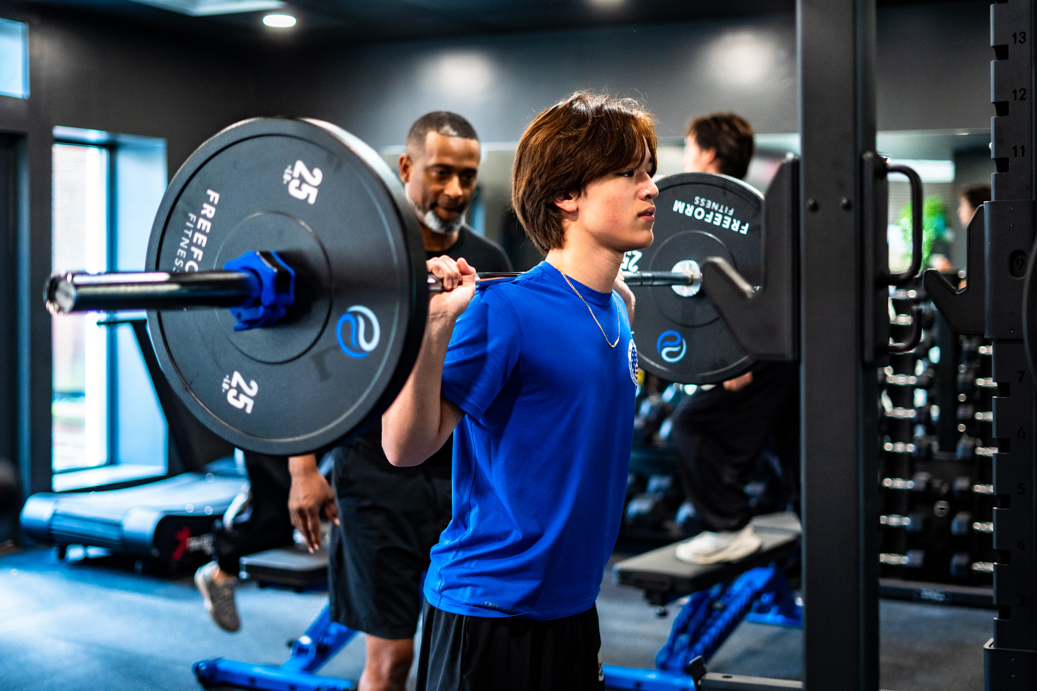 Young man in blue shirt lifting a barbell in a gym, with two people in the background. Bright gym environment.
