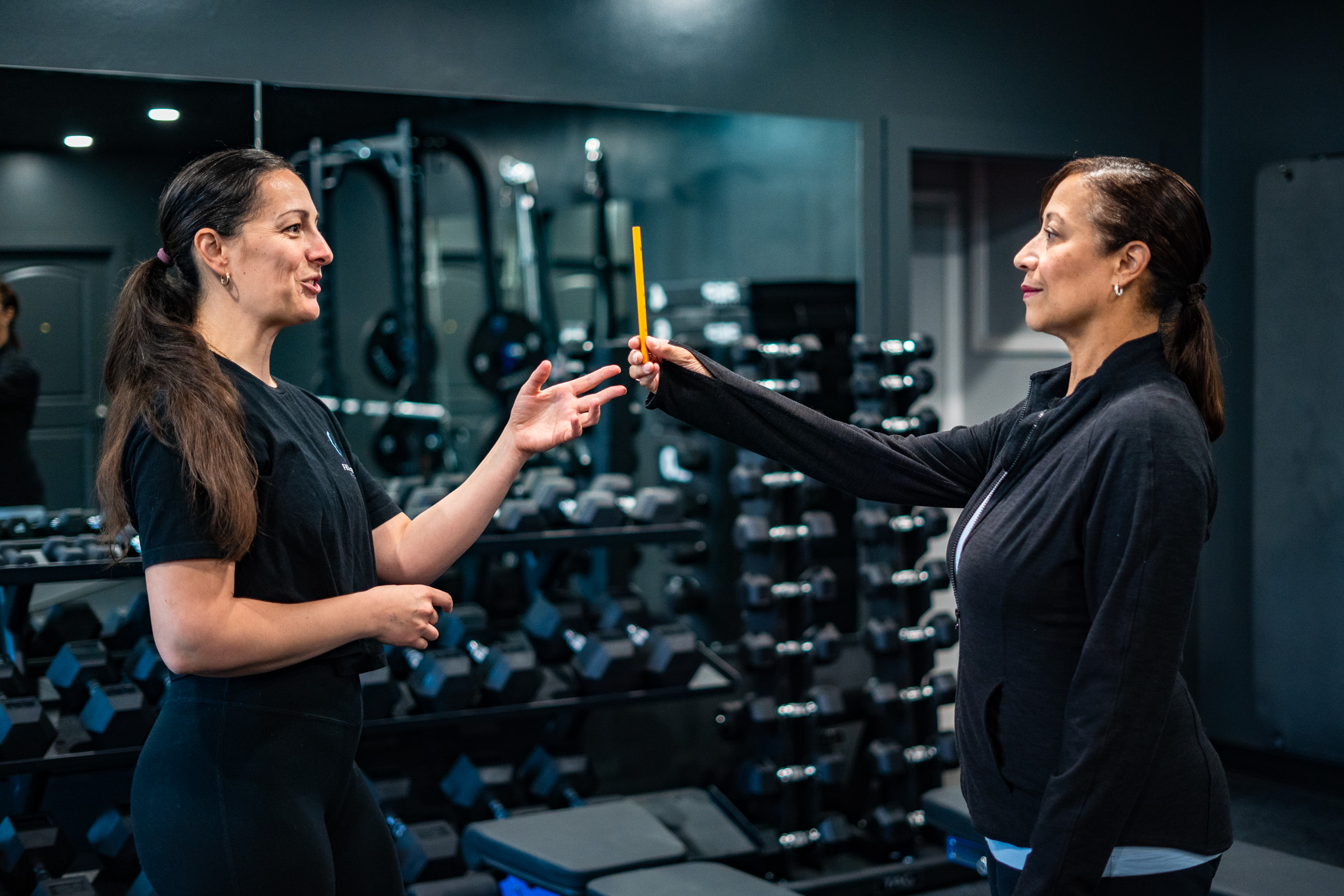 Two women exchanging a yellow object in a gym with exercise equipment in the background.