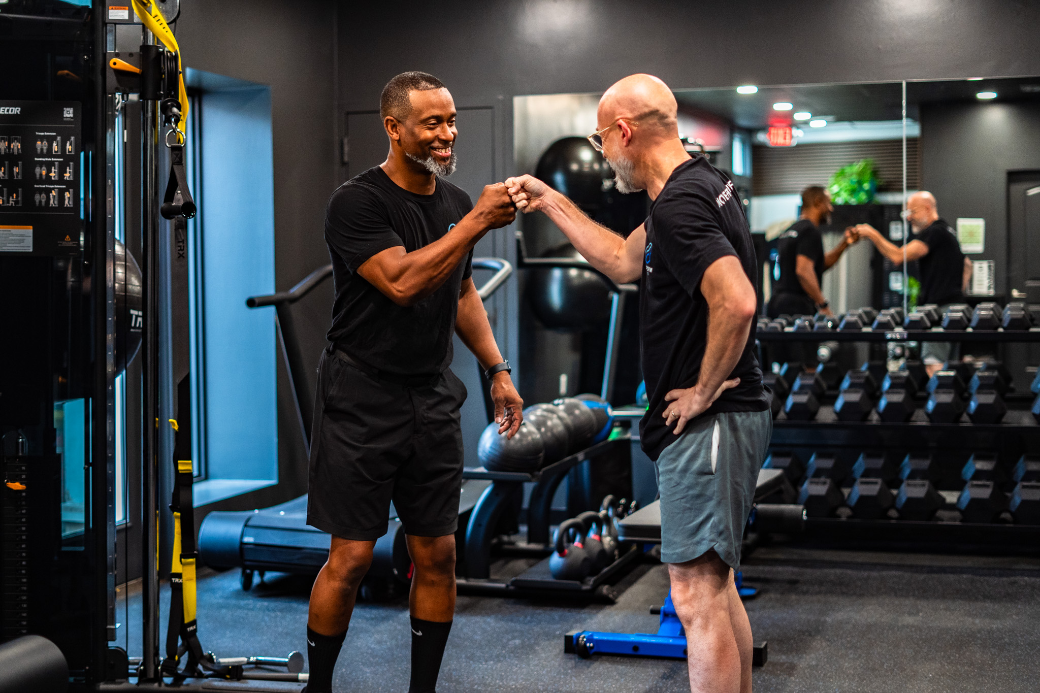 Two men shake hands in a gym, surrounded by exercise equipment and mirrors, smiling at each other.