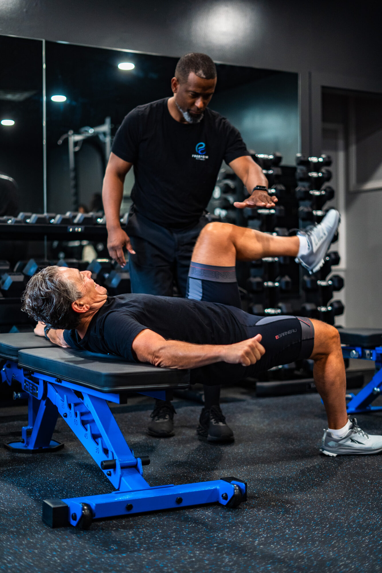 A woman lying on a workout bench with a trainer assisting her leg exercise in a gym. Equipment visible in background.