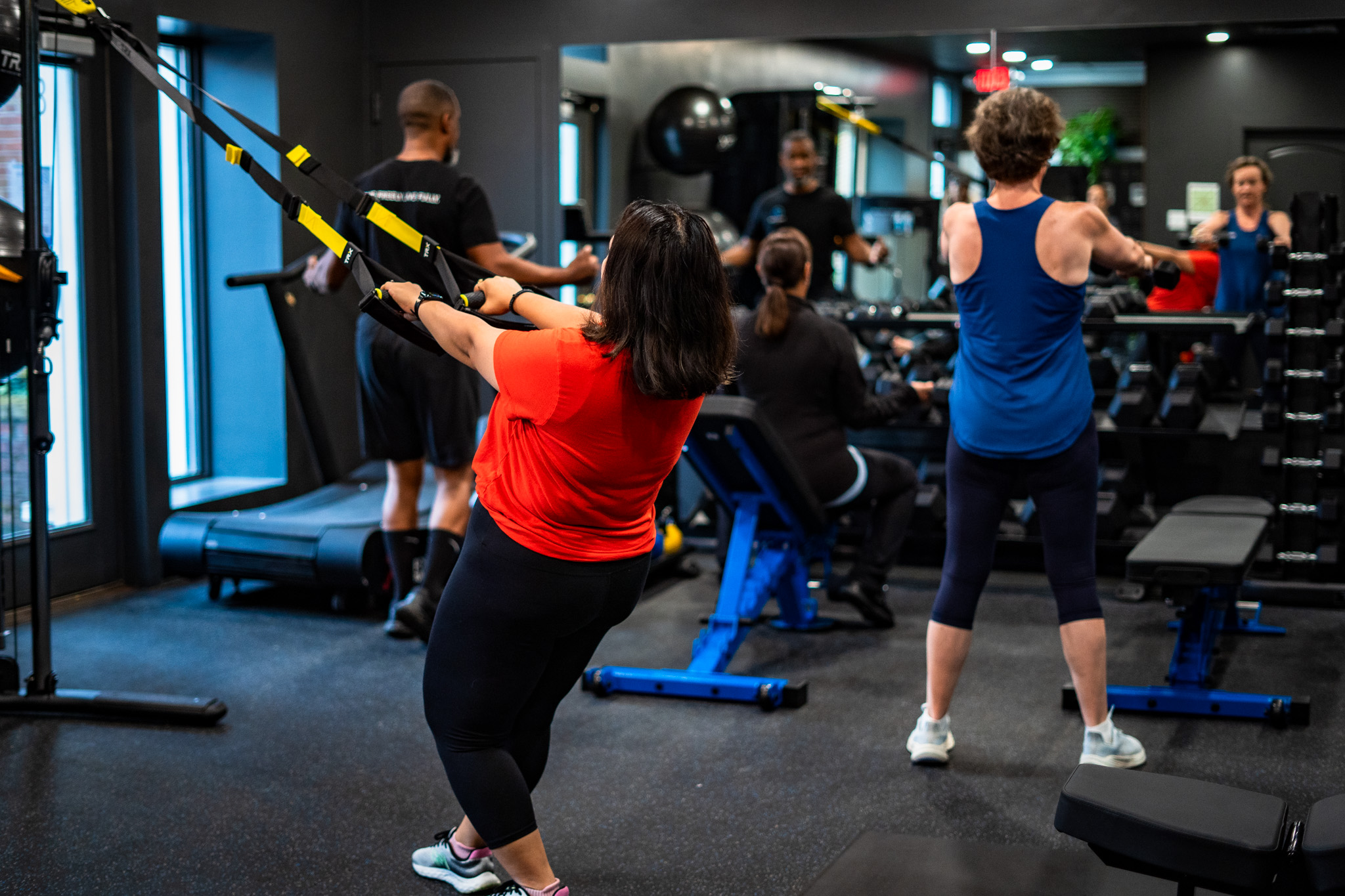 People exercising with resistance bands and weights in a gym, some standing and some seated, with gym equipment visible.