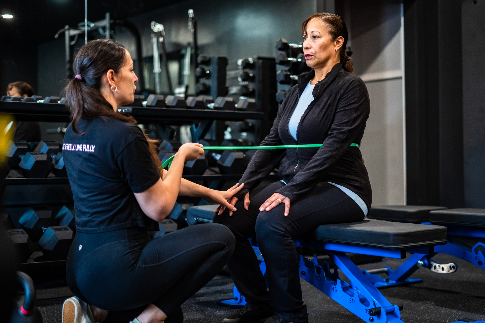 A woman in workout attire sits on a bench while a trainer measures her waist with a green tape measure in a gym.