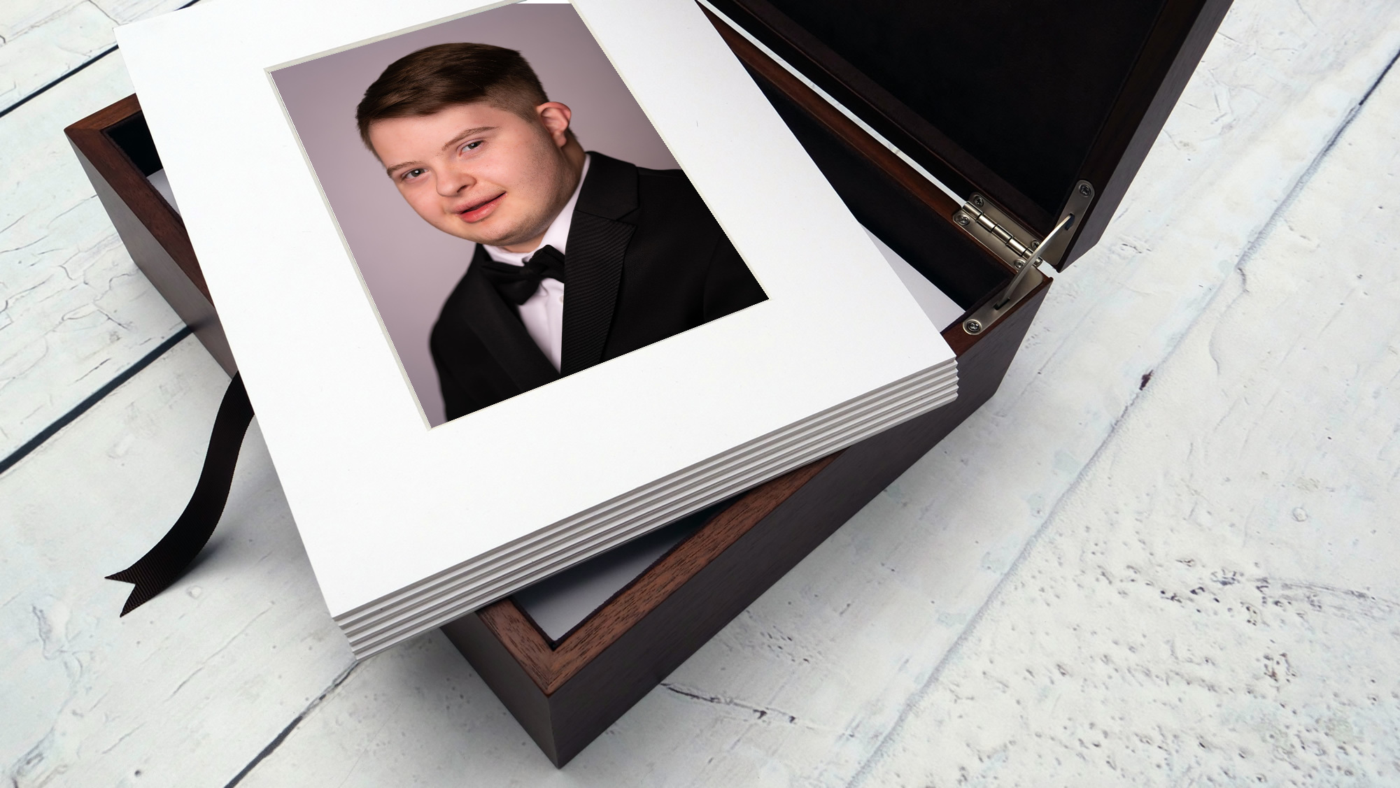 Stack of photo prints with a young boy in a tuxedo on top, inside a wooden box, on a white surface.