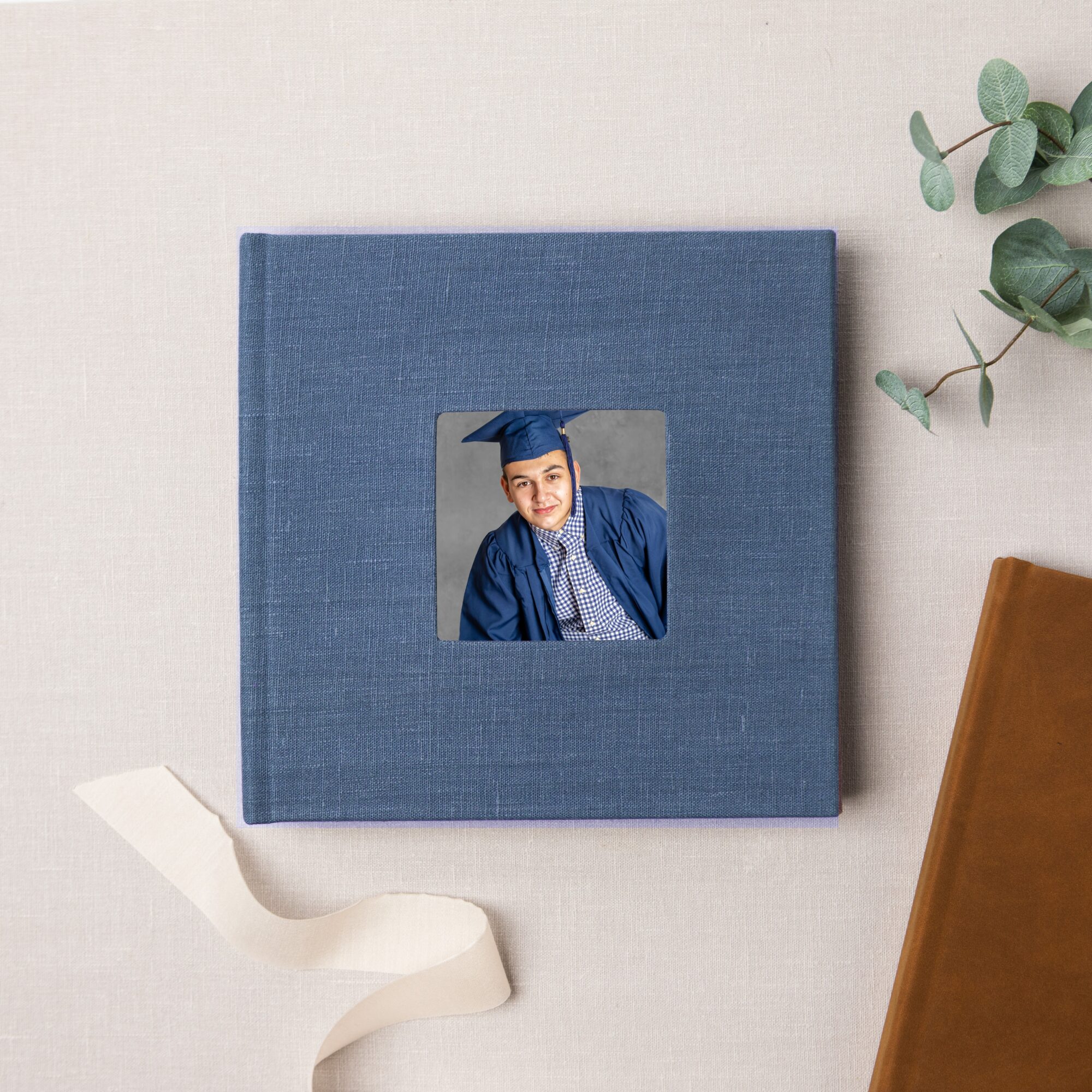 Blue graduation photo album with a picture of a graduate in cap and gown, on a light surface with green leaves and a brown object nearby.
