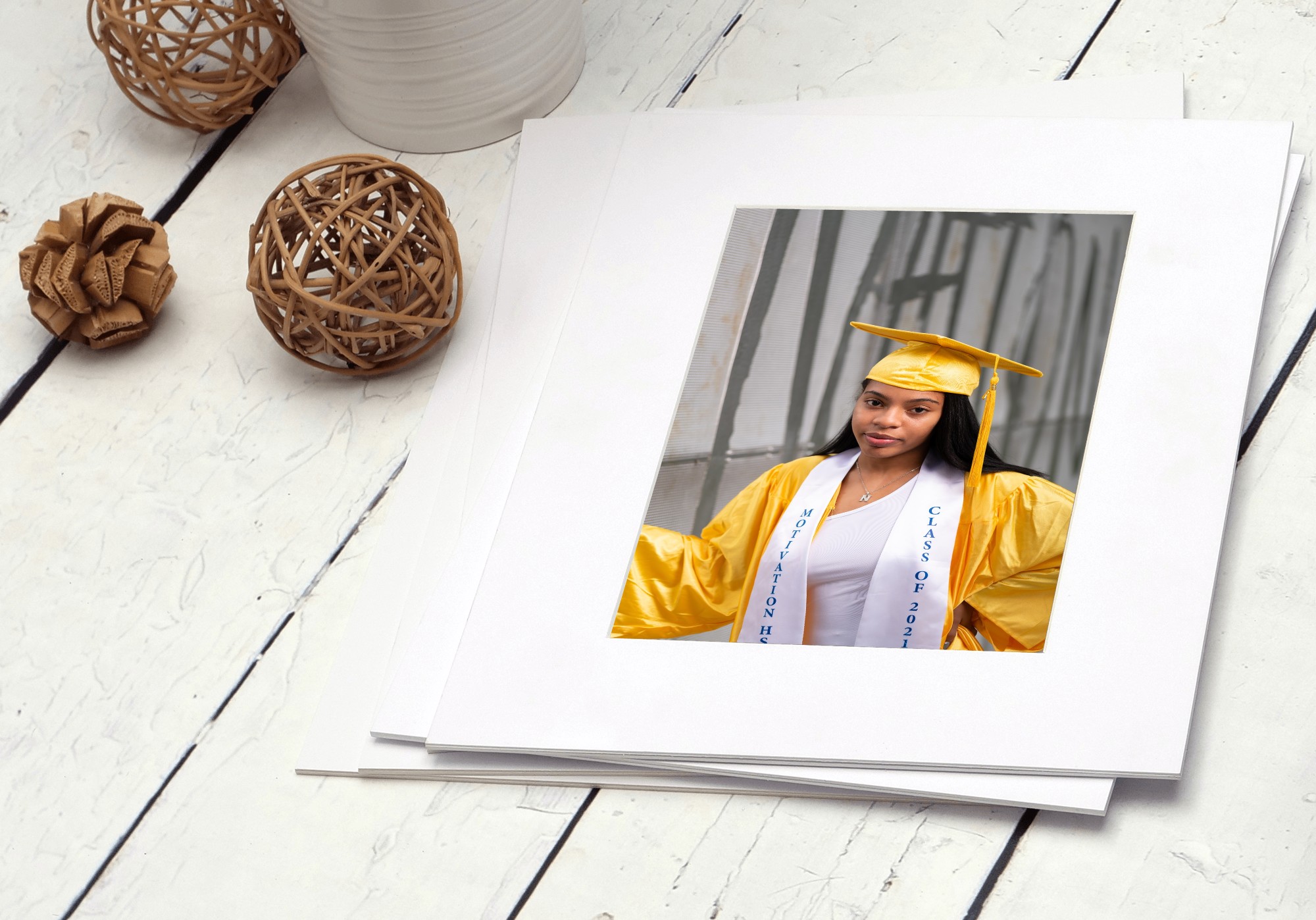 Graduation photo of a young woman in a yellow cap and gown, holding a diploma, on a white surface with decorative objects.