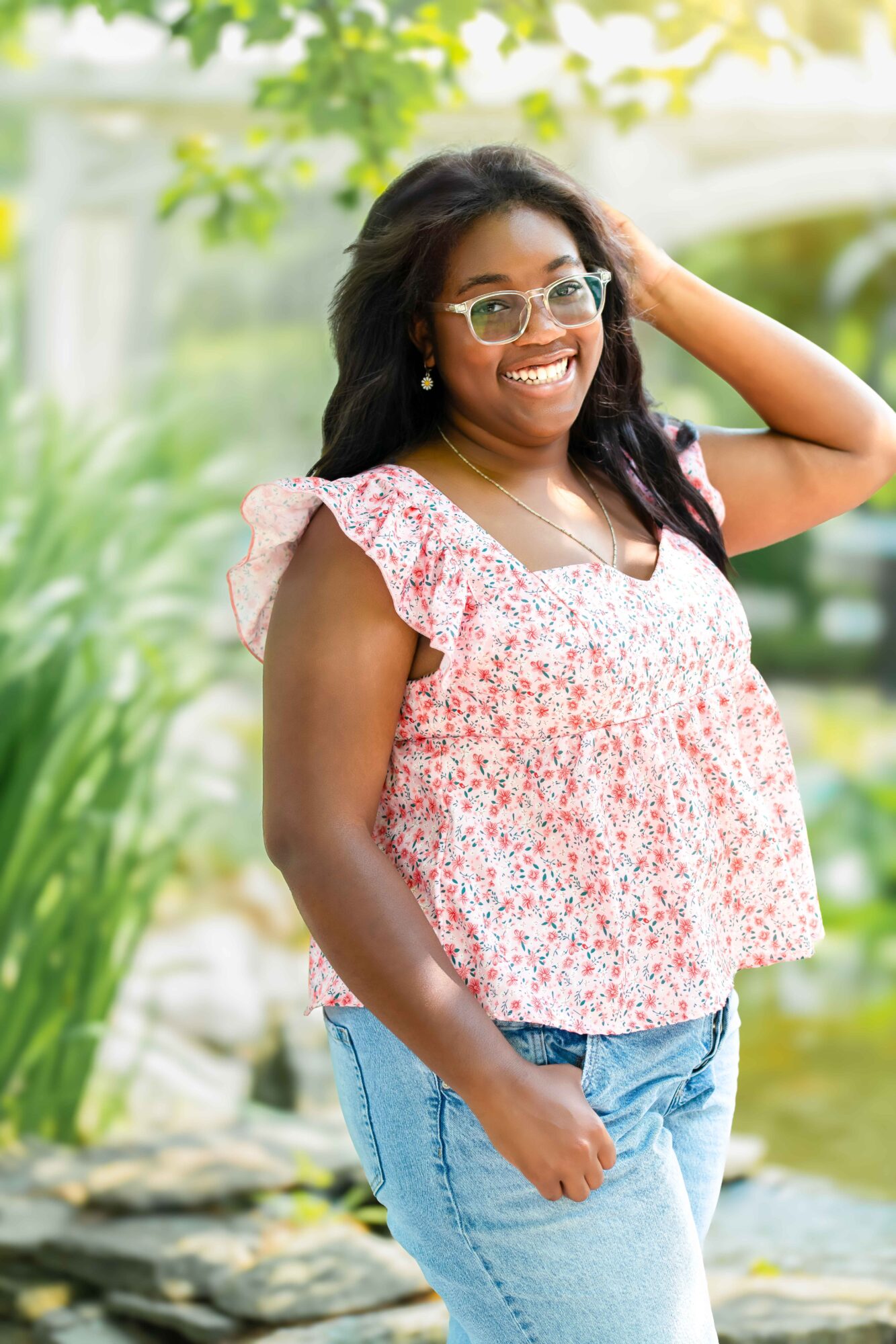 Smiling woman with glasses and long dark hair outdoors, wearing a pink floral top and blue jeans, standing near water.