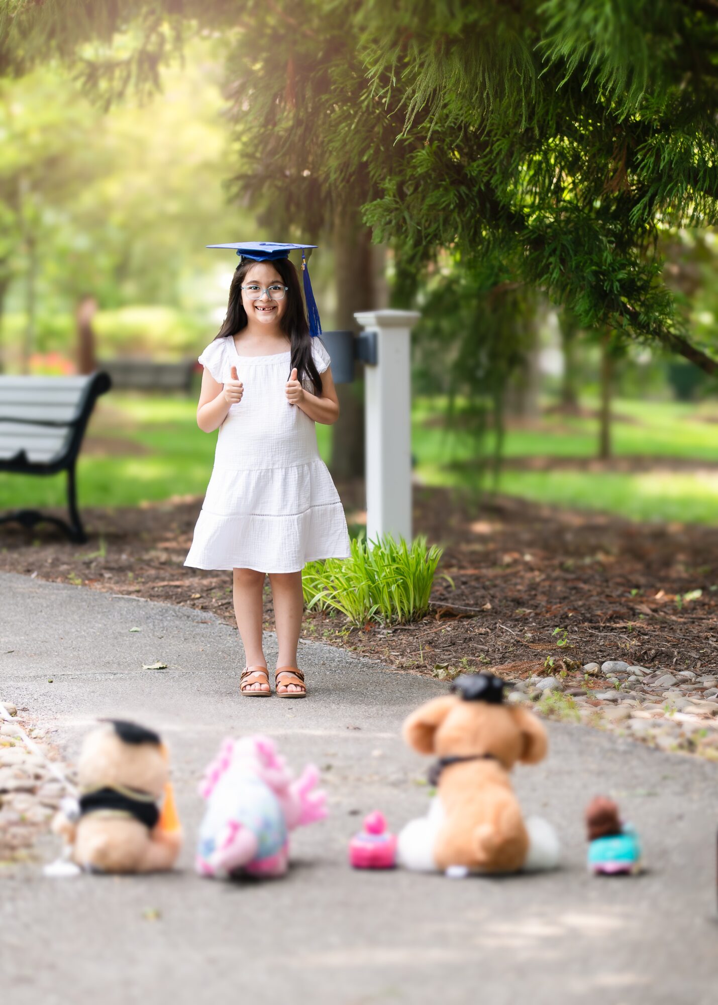 Young girl in a white dress with a graduation cap, standing outdoors near a park bench and trees, with stuffed animals on the ground.