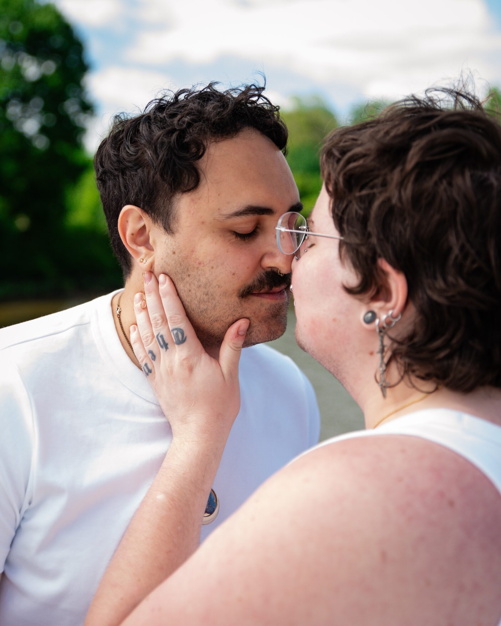 Two people face each other outdoors, about to kiss, with trees and sky in the background.