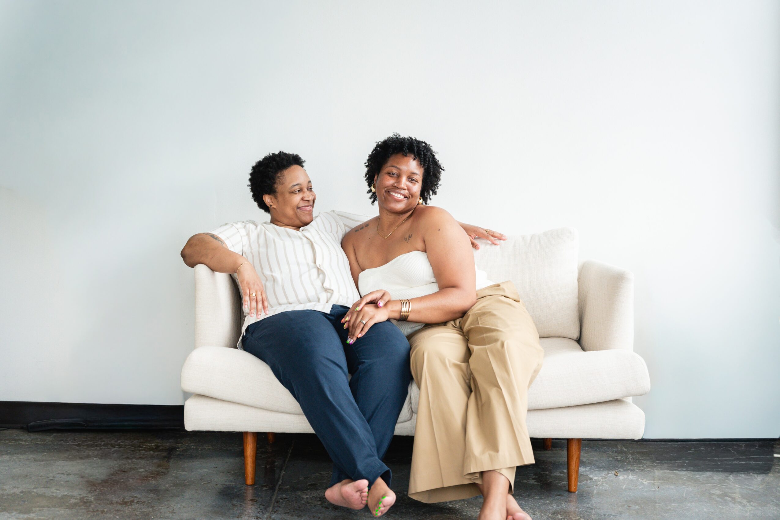 Two women sitting on a white sofa, smiling and laughing, against a plain light-colored wall.