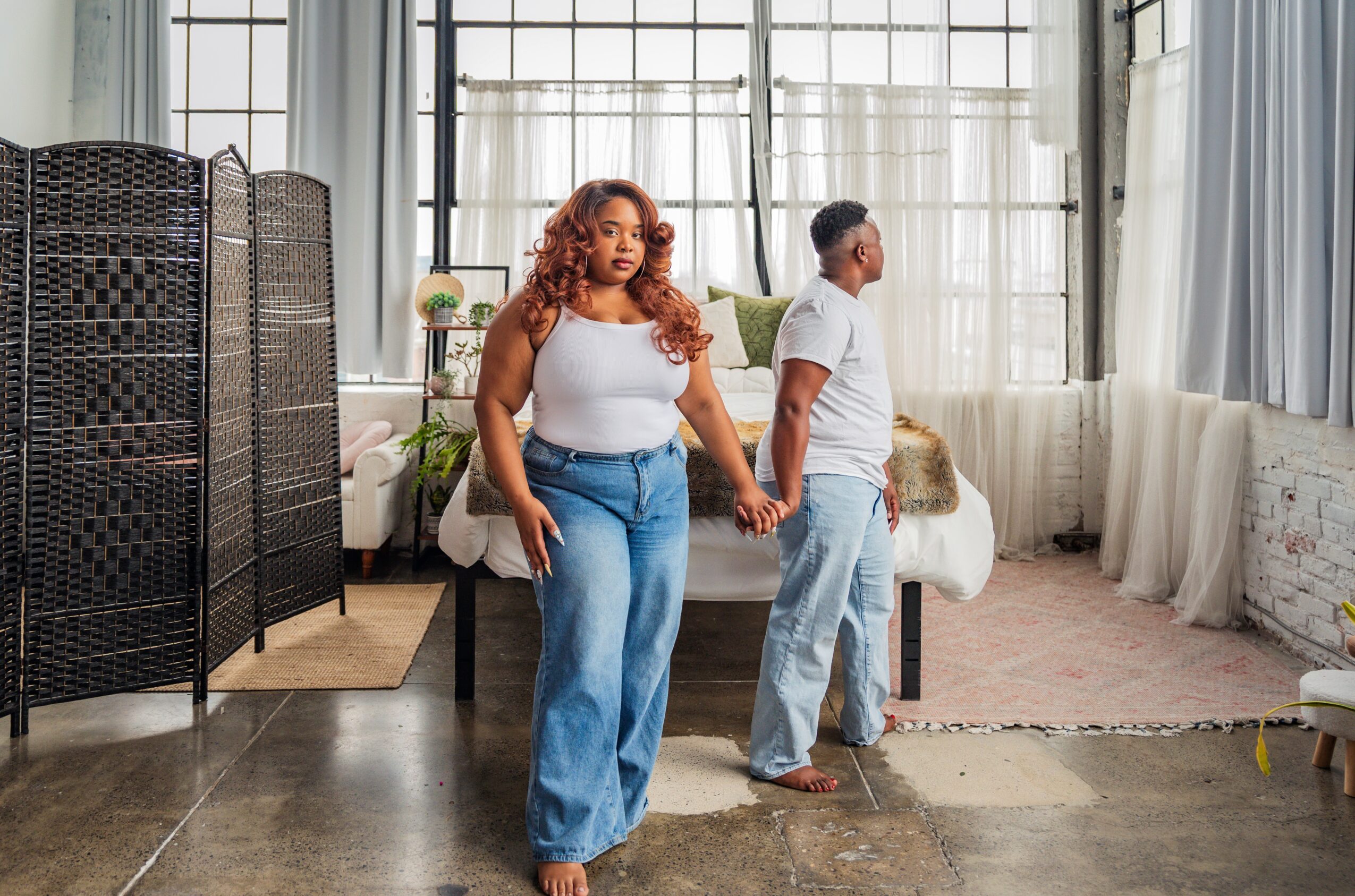 Two women stand in a bright room with large windows, one facing forward and the other turned away, holding hands.
