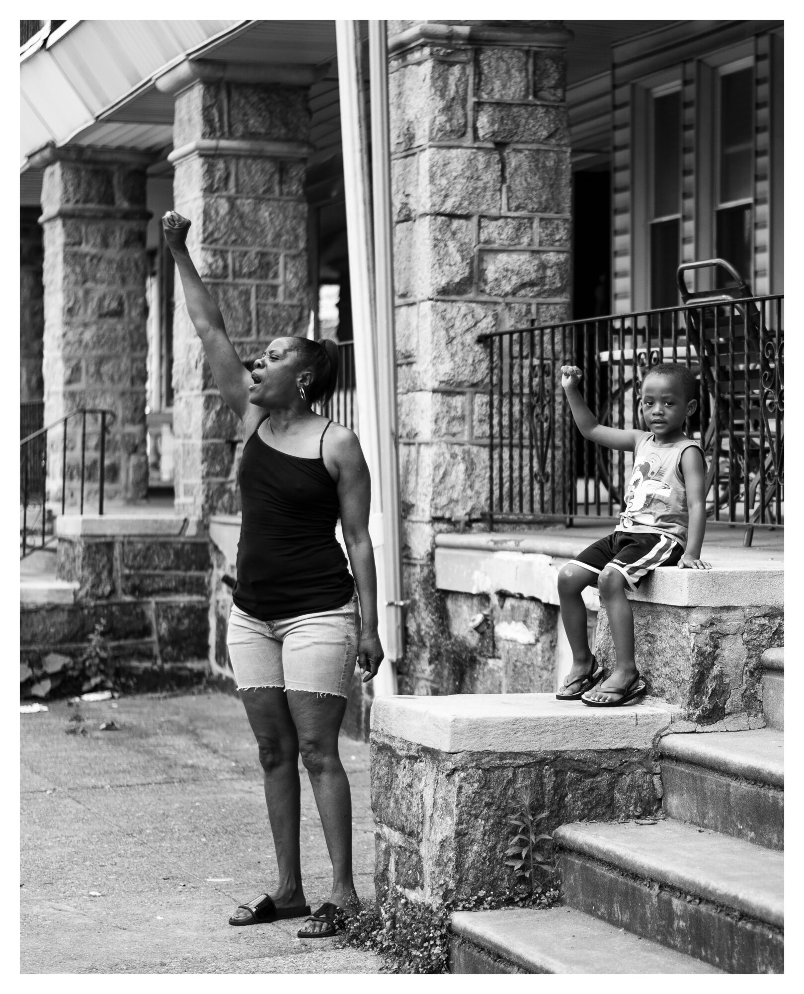 A woman and a child sit on stone steps outside a building with stone columns and a metal railing, in black and white.