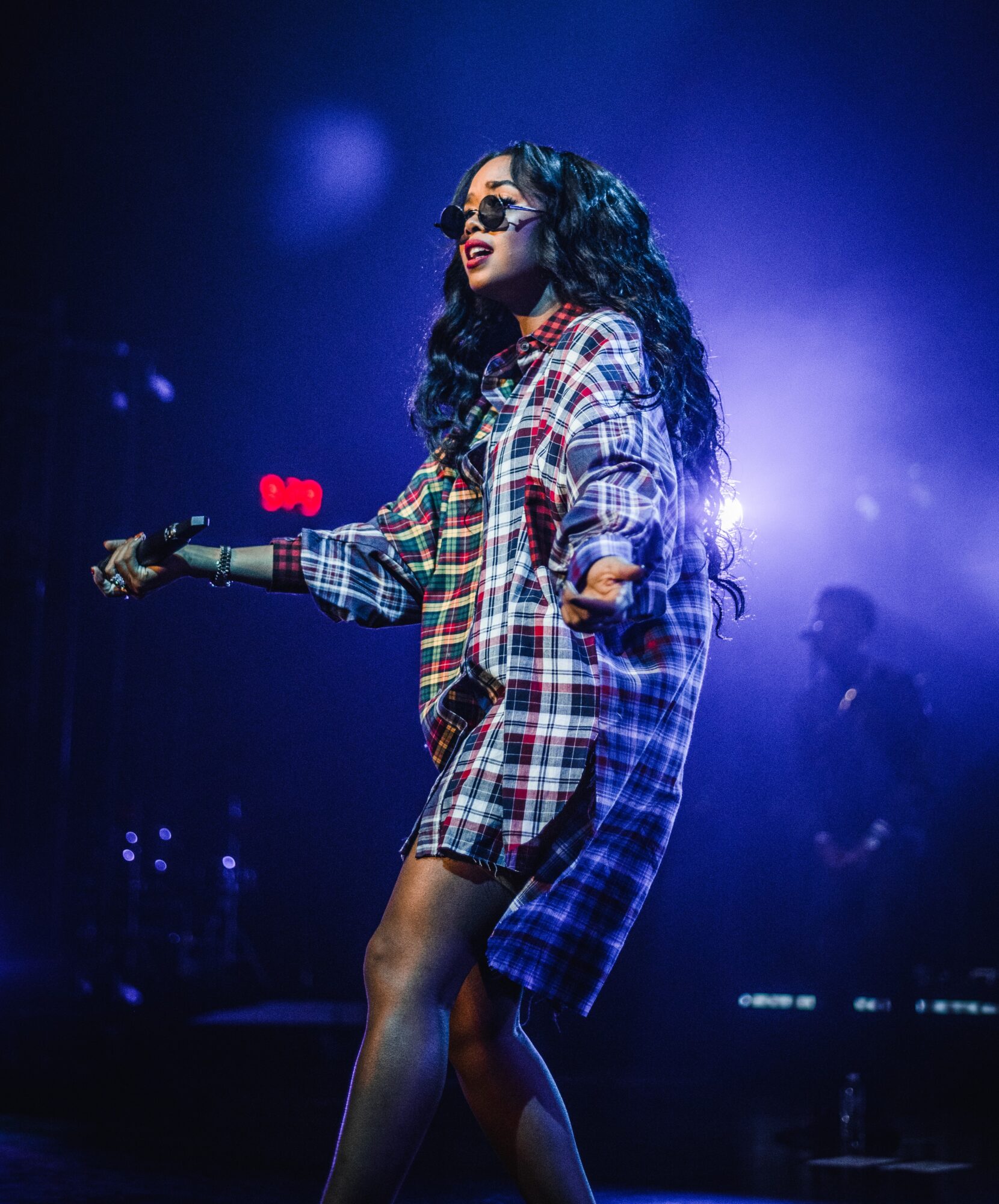 A woman with long curly hair singing on stage, holding a microphone, wearing a plaid shirt, with blue and purple stage lighting.