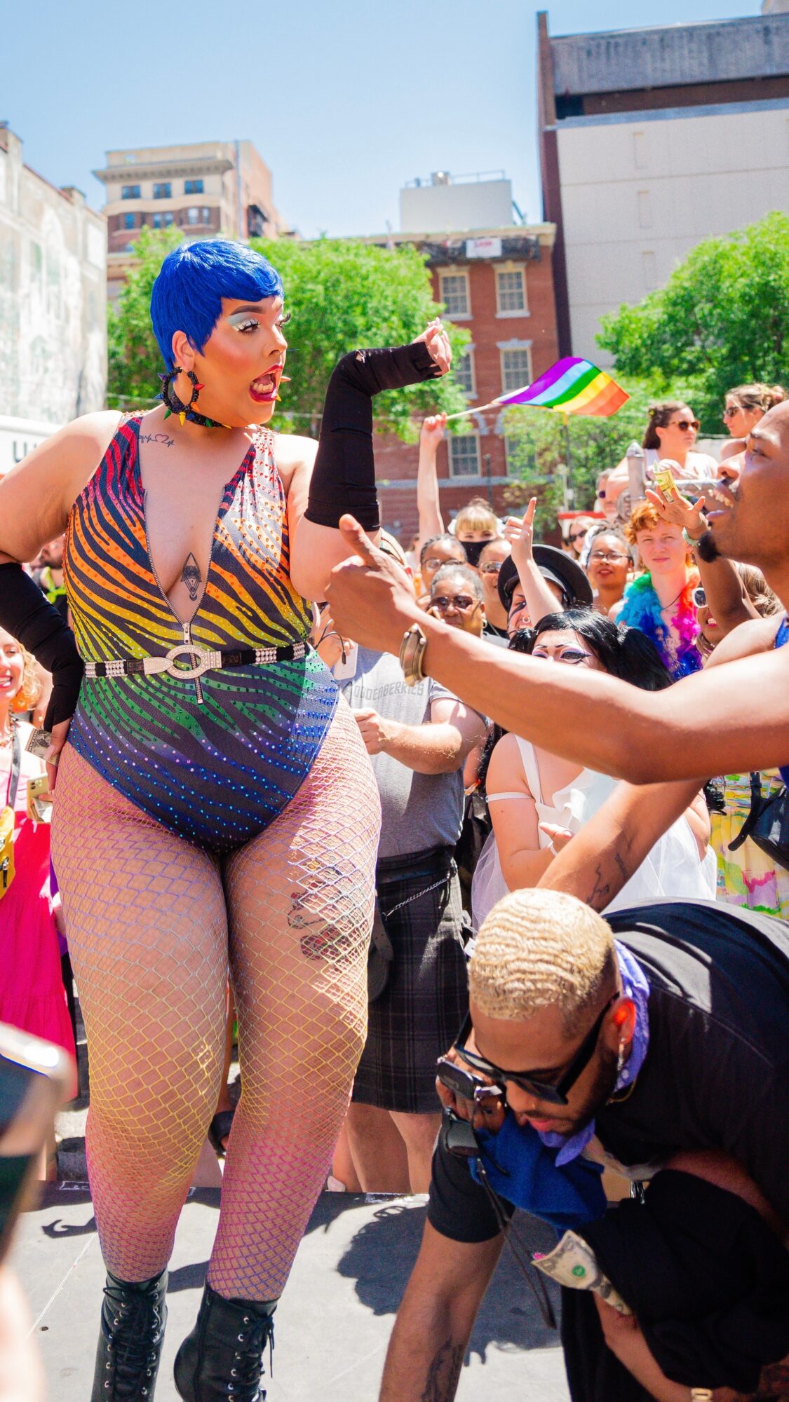 Person with blue hair and colorful outfit interacting with crowd outdoors during daytime, with buildings and trees in background.