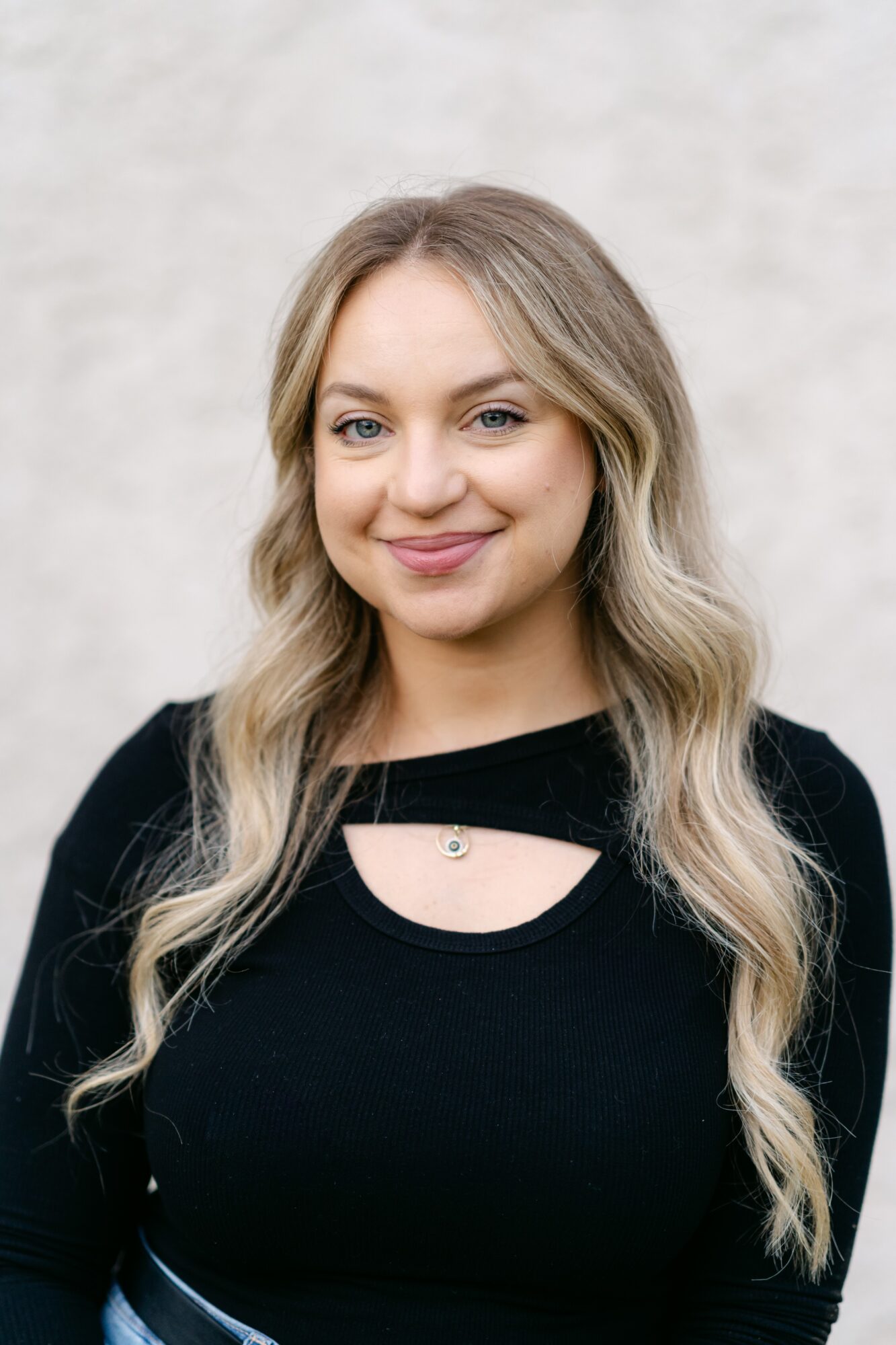 Young woman with long wavy blonde hair smiling, wearing a black top with a cutout, standing against a light background.