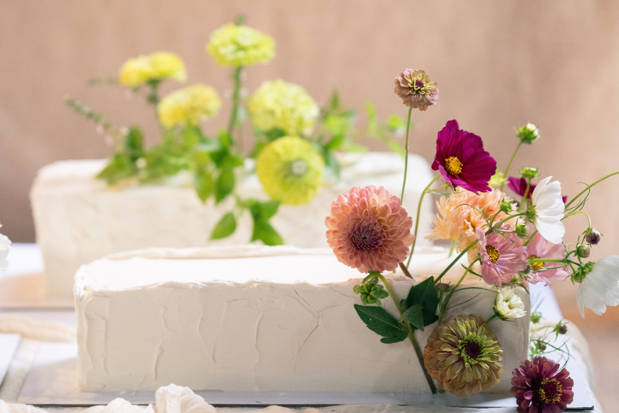 Two white cakes decorated with colorful flowers, with a blurred background of more flowers. Word count: 20.