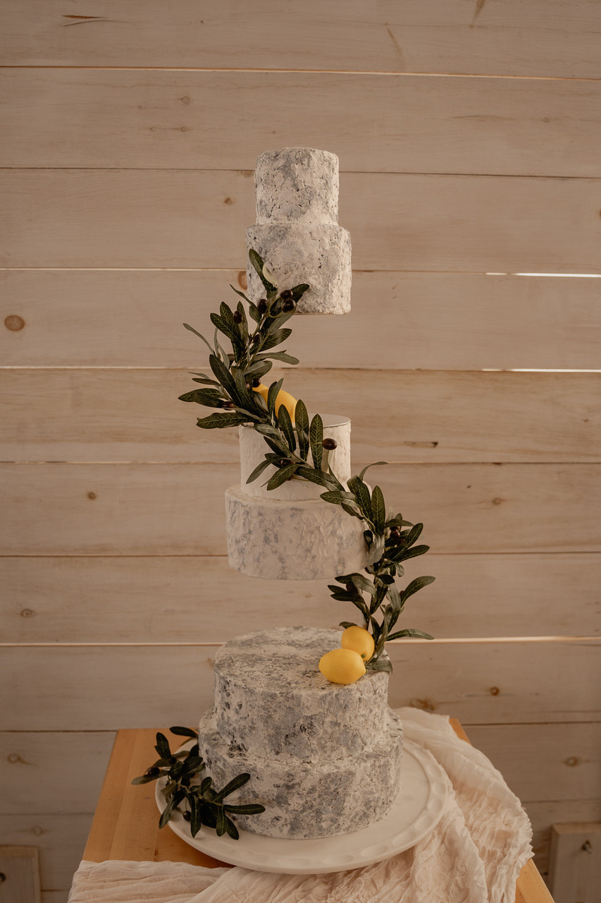 A rustic, two-tiered cake with a branch of greenery and lemons, set on a wooden surface against a wooden wall.