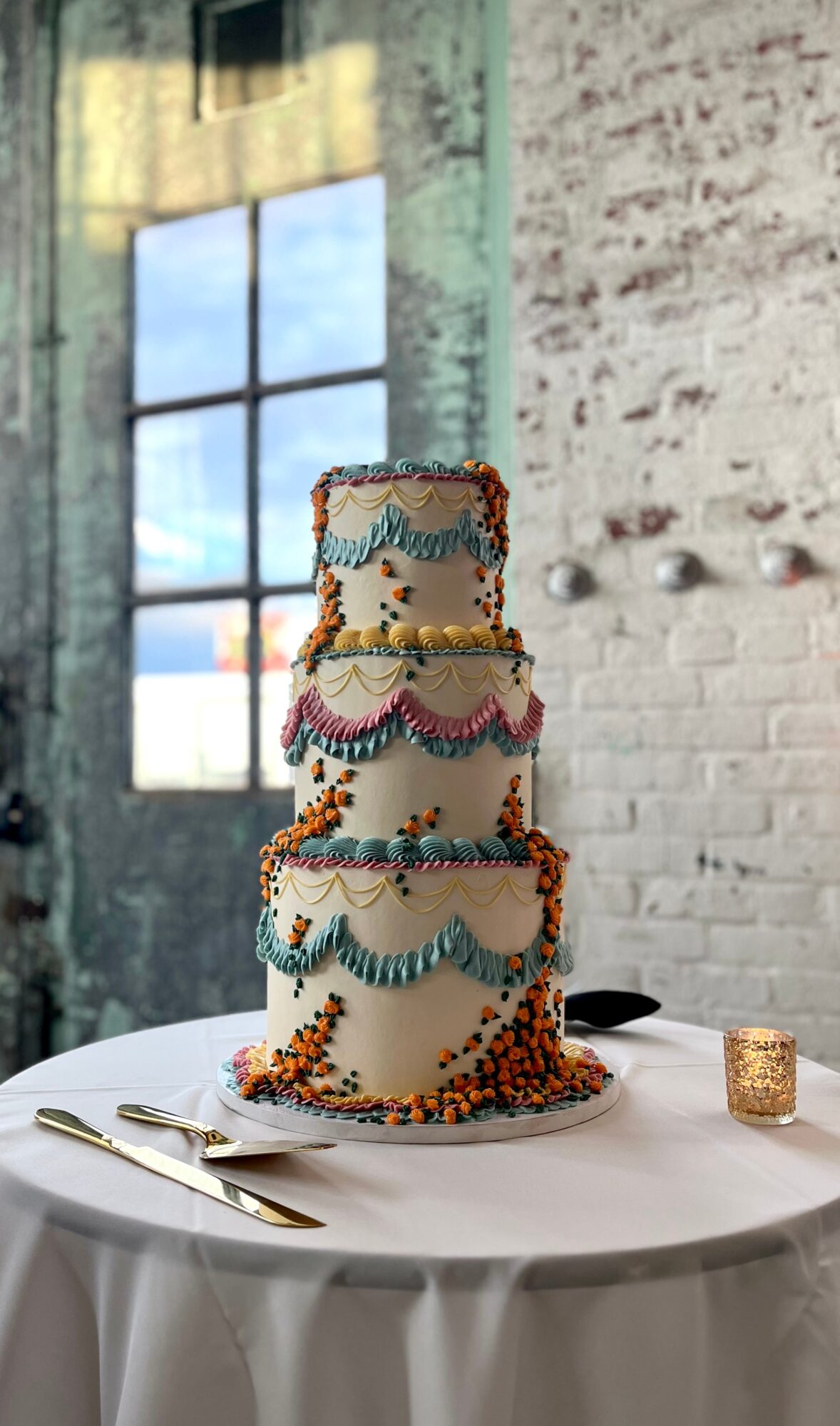 Three-tiered cake decorated with colorful icing and sprinkles, on a white table with a gold candle holder and utensils, in a rustic room.
