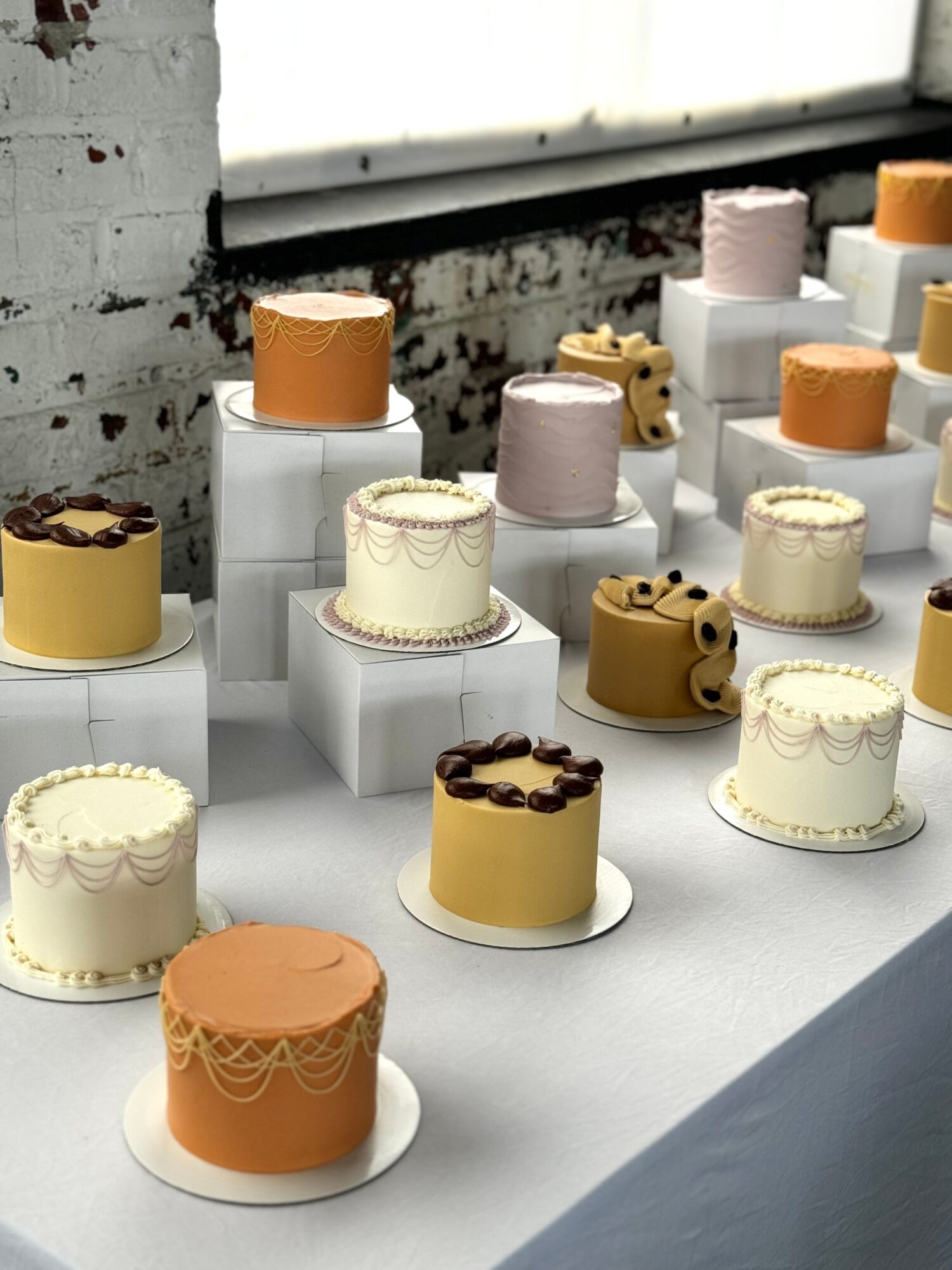Display of various decorated cakes on white stands, with a brick wall and window in the background.