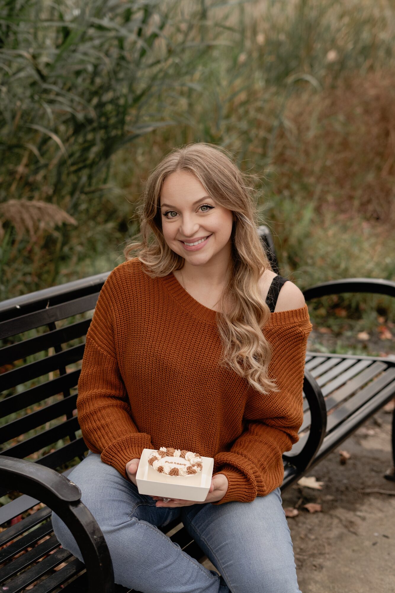 Young woman with long wavy hair holding a cake, sitting on a park bench outdoors, smiling.