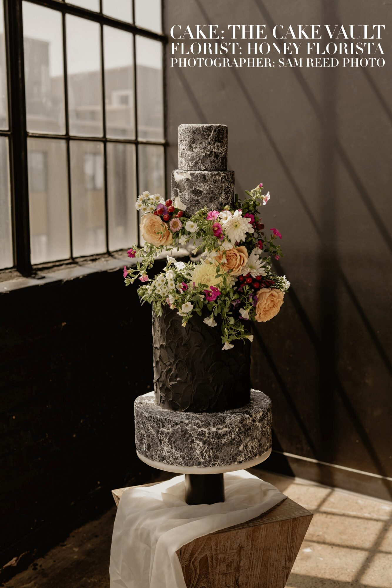 Three-tiered cake with floral decorations on a stand, sunlight streaming through window, shadows on wall.