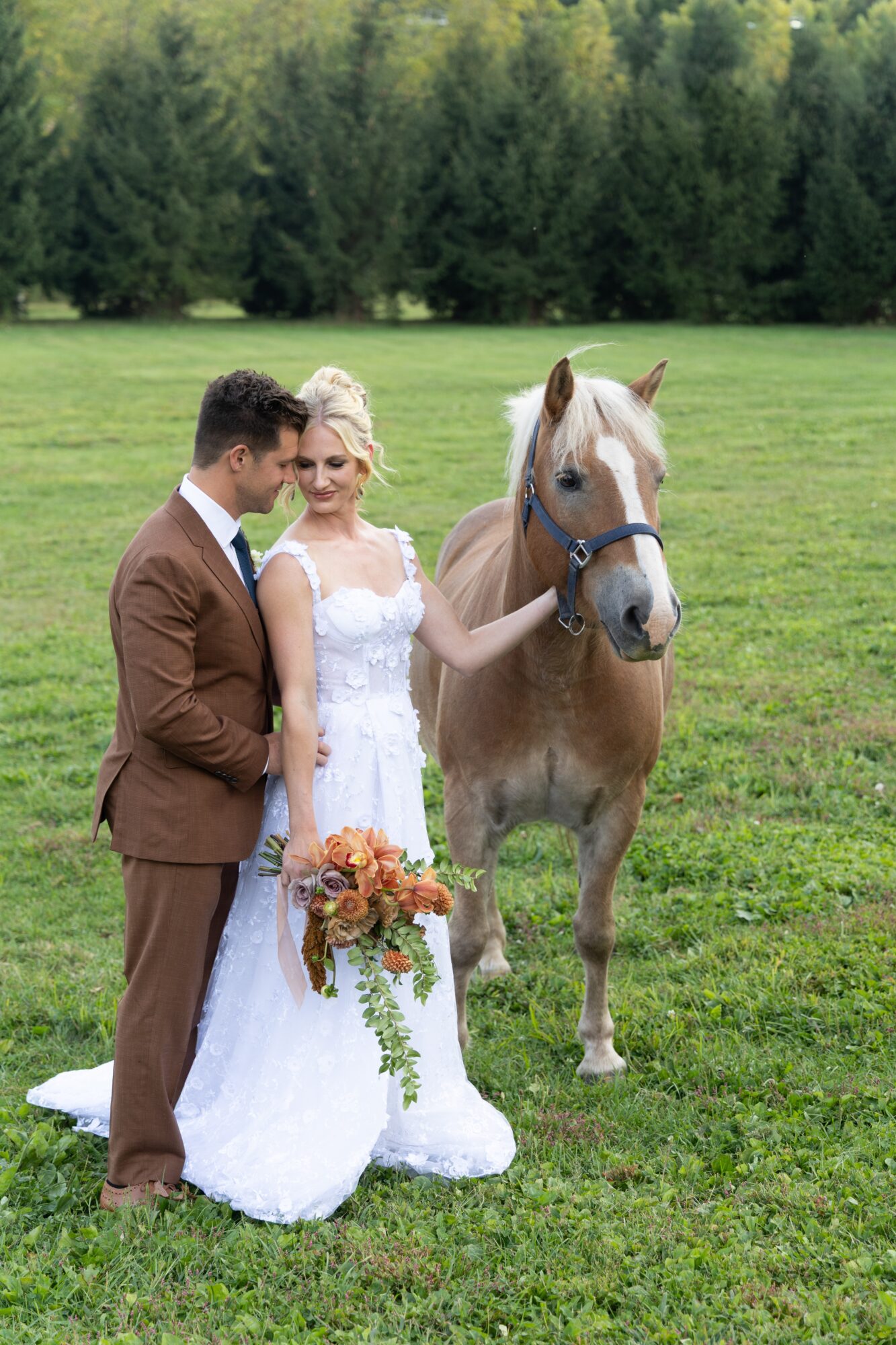 Bride and groom stand on grass with a horse, bride holding a bouquet, in a green outdoor setting.