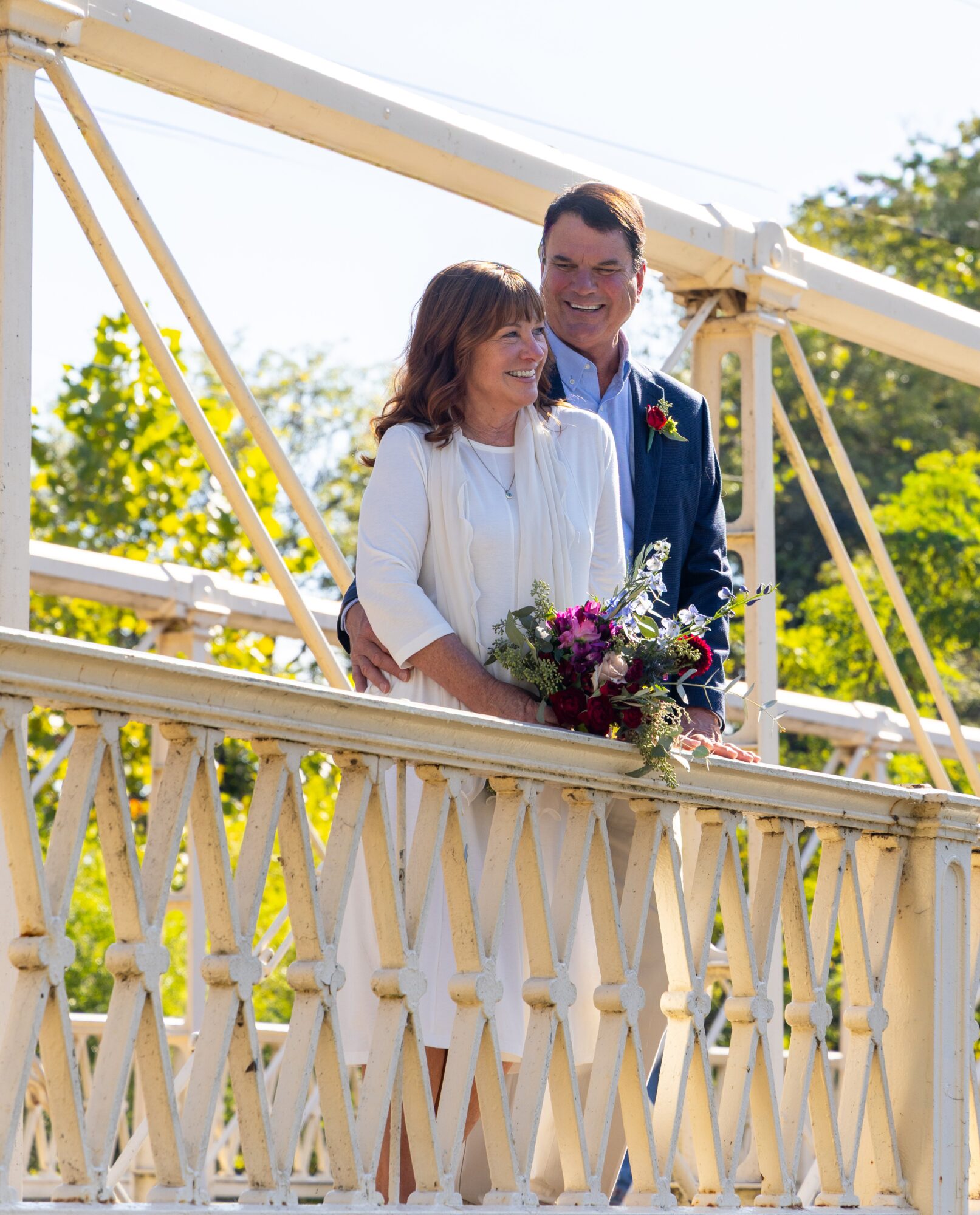 A couple stands on a bridge, smiling, with trees and blue sky in the background, holding a bouquet of flowers.