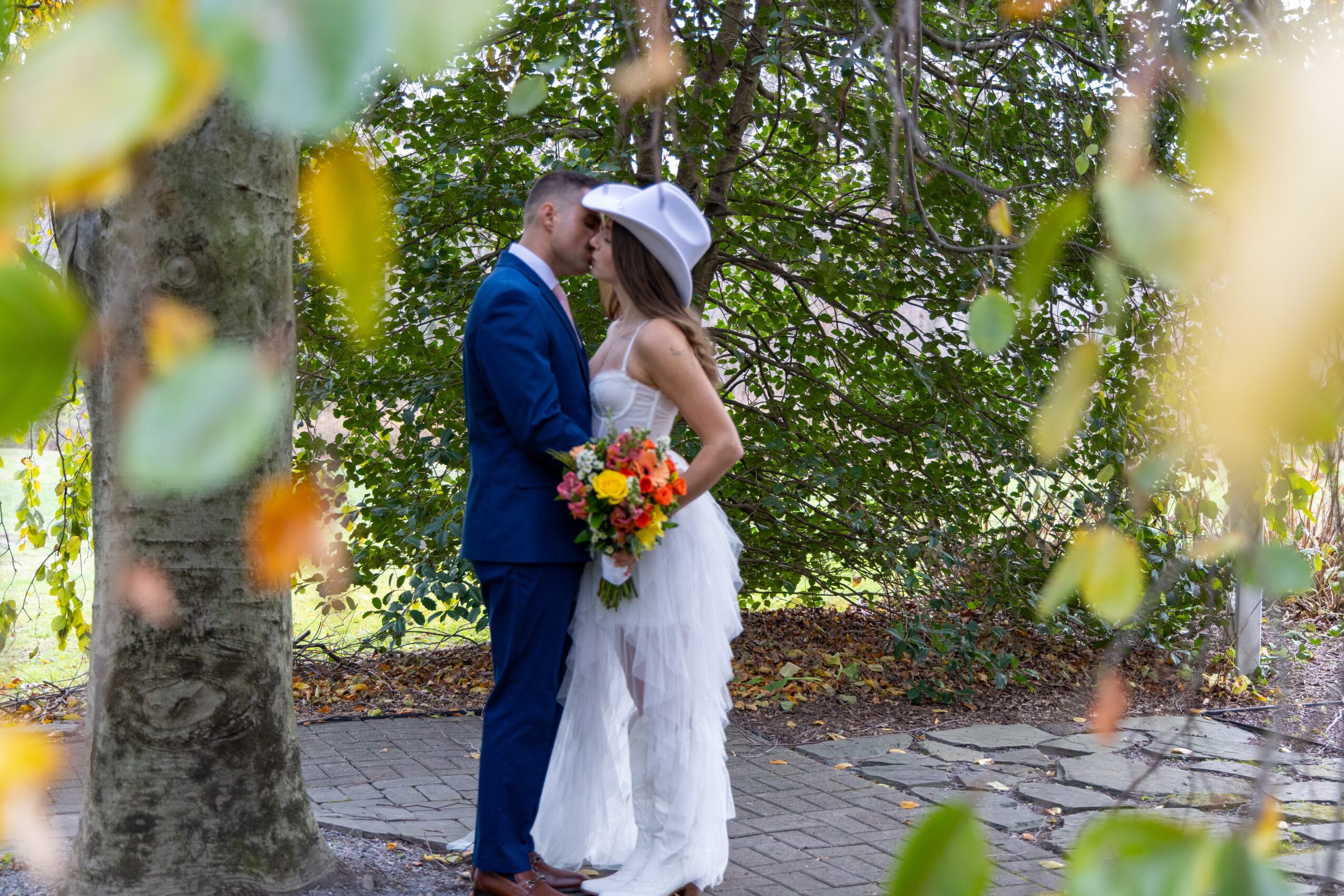 Bride and groom stand close together outdoors, surrounded by trees, with the bride holding a colorful bouquet, both in wedding attire.