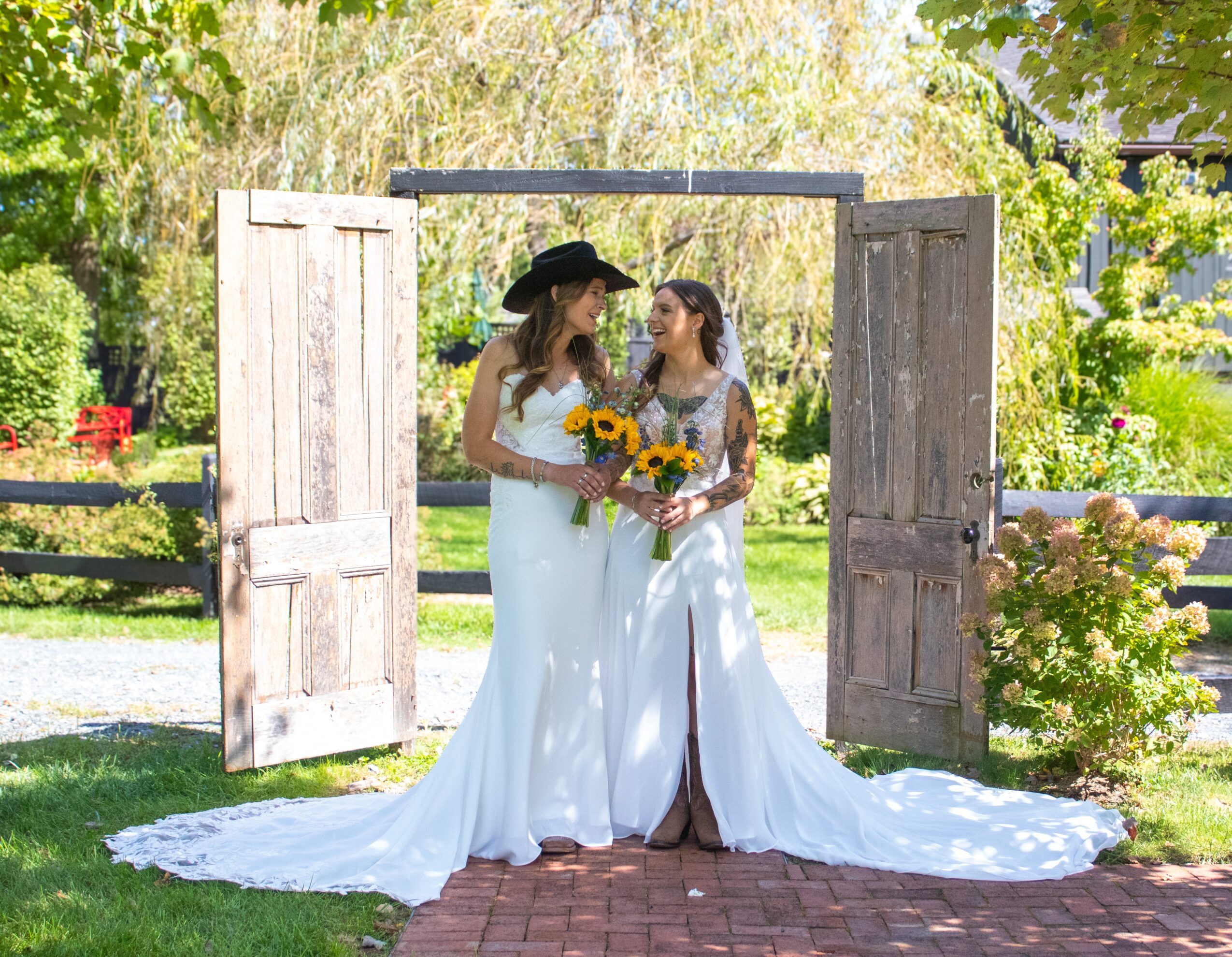 Two women in white wedding dresses stand outdoors in front of open wooden doors, holding a bouquet of sunflowers, smiling at each other.