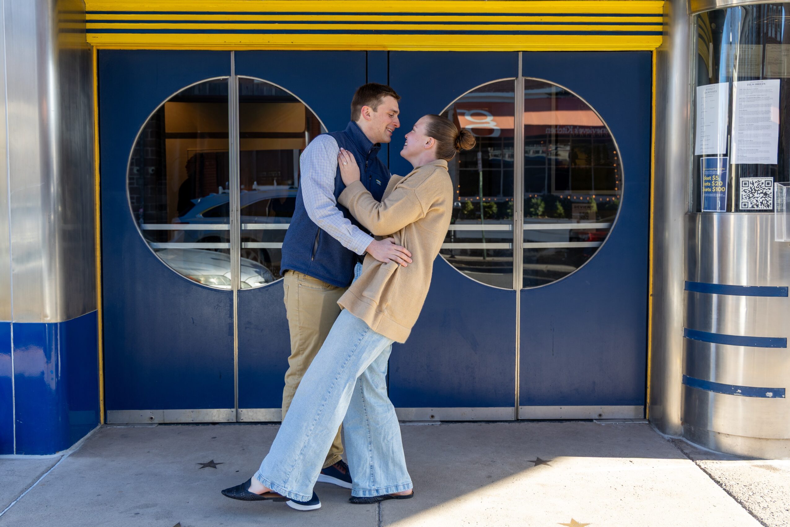 A couple stands close, facing each other, in front of a blue door with circular windows, outside a building.