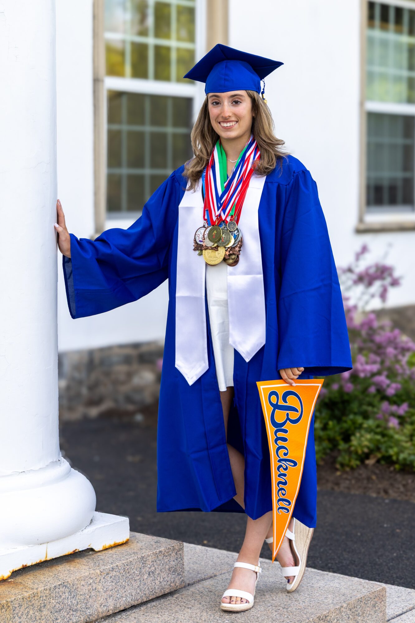 Young woman in blue graduation gown and cap holding a diploma, wearing multiple medals, standing outside near a white column.