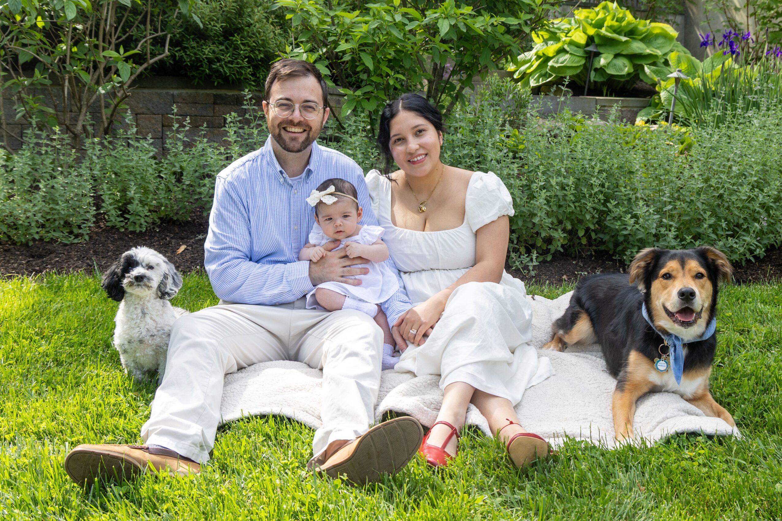 Family of three with two dogs sitting on grass in a garden, smiling, with greenery in the background.