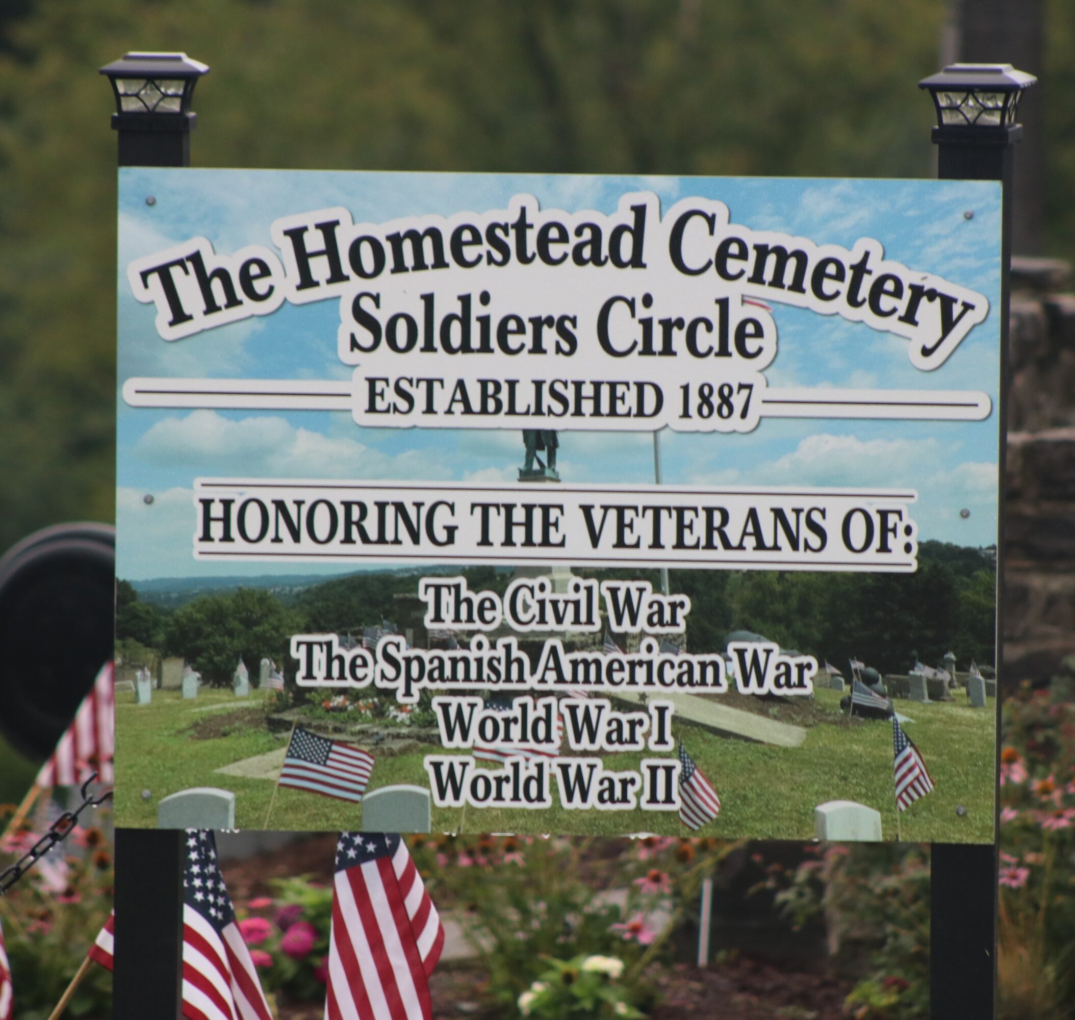 Sign for Homestead Cemetery Soldiers Circle honoring veterans of wars, established 1887, with flags and graves in background.