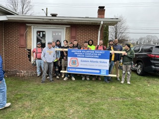 Group of people holding a large banner outside a brick house with a lawn and parked cars nearby.