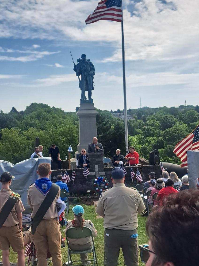 People gather at a monument with a soldier statue, American flags, and a cloudy sky background.