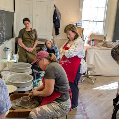 People working on pottery in a classroom with a teacher observing, others watching, and sunlight coming through windows.