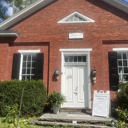 Brick house with white door, black shutters, and a sign outside, surrounded by greenery and potted plants.