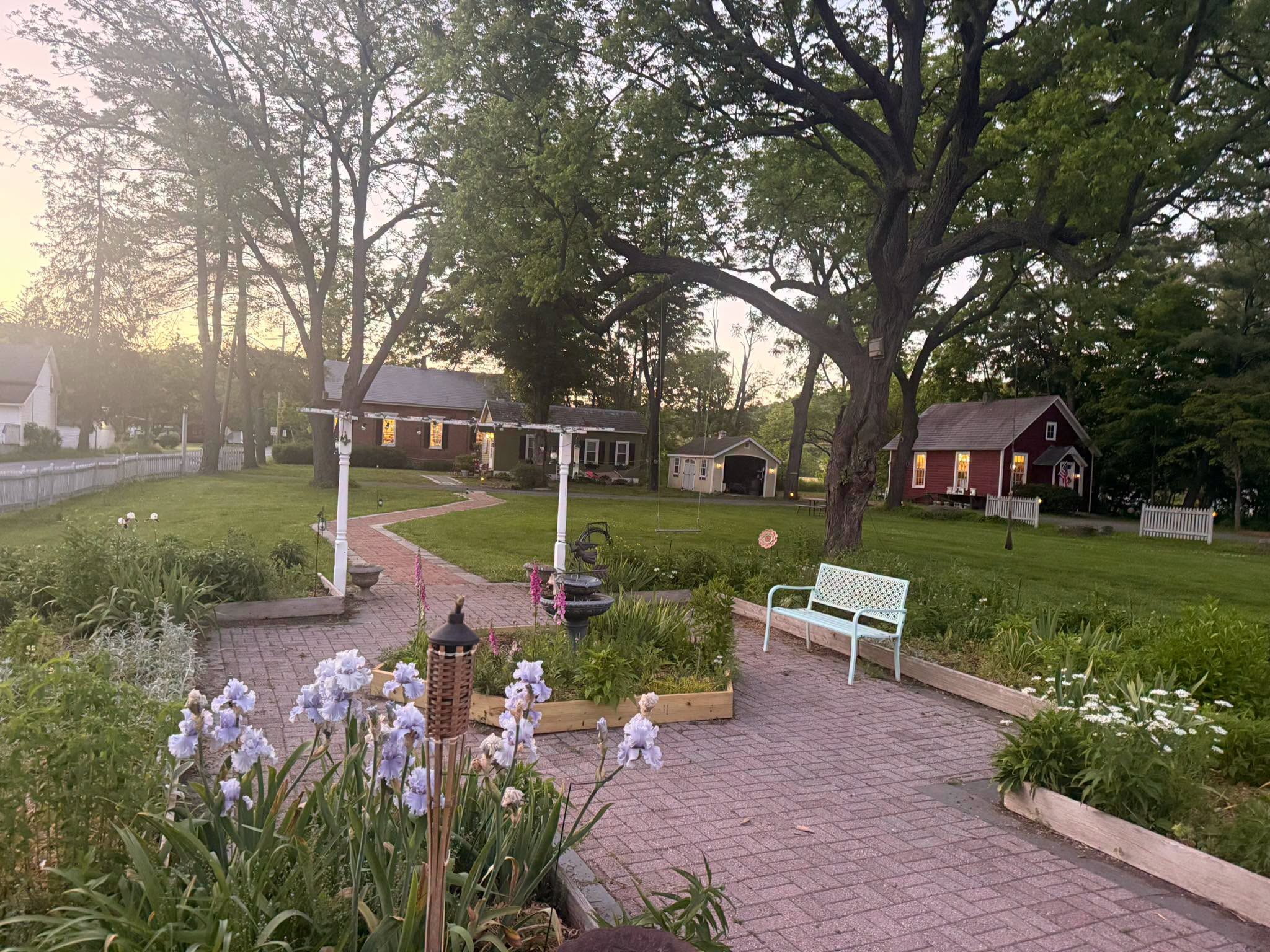 Park with trees, brick pathway, white bench, and flowers, buildings in background, sunlight filtering through trees.